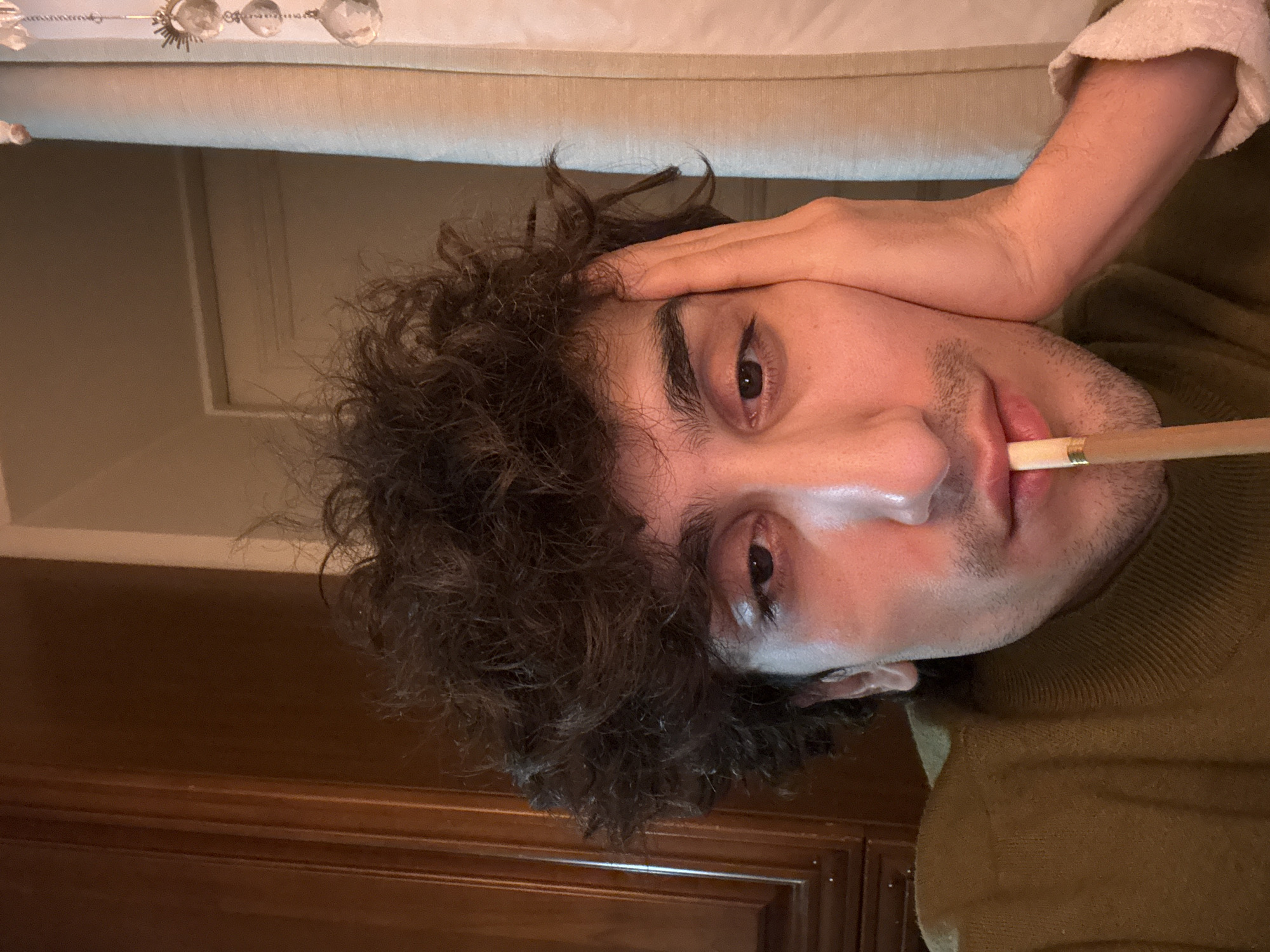 A man with dark curly hair holds a thin, light brown object in his mouth. He is wearing a dark green turtleneck sweater. His hand is touching his forehead. A wooden cabinet is visible in the background, as well as a beige upholstered piece of furniture. There is no indication of the location being Moscow, Russia.