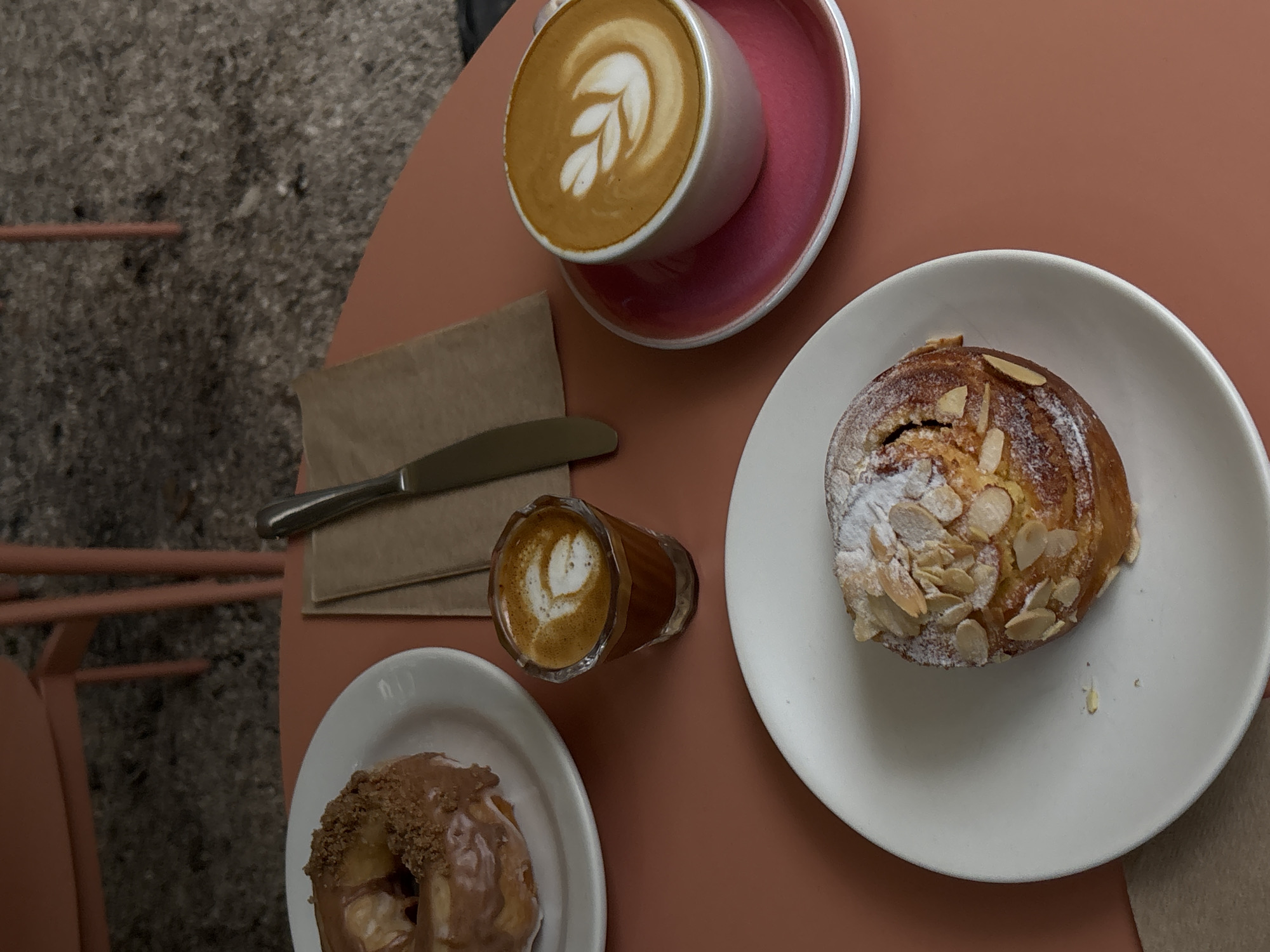 On a terracotta-colored table, viewed from directly above, are an assortment of pastries and coffee beverages. The scene includes a cappuccino with latte art in a white mug with a rose-colored saucer, a small glass of espresso with latte art, and two pastries served on separate white plates. A metal butter knife rests on a folded brown paper napkin. The pastries are different types; one is sprinkled with almond slivers and powdered sugar, and the other is glazed with a dark topping and appears to have a crumbled topping. The setting suggests a cafe or coffee shop, but the specific location in Unknown, Unknown is not discernible.