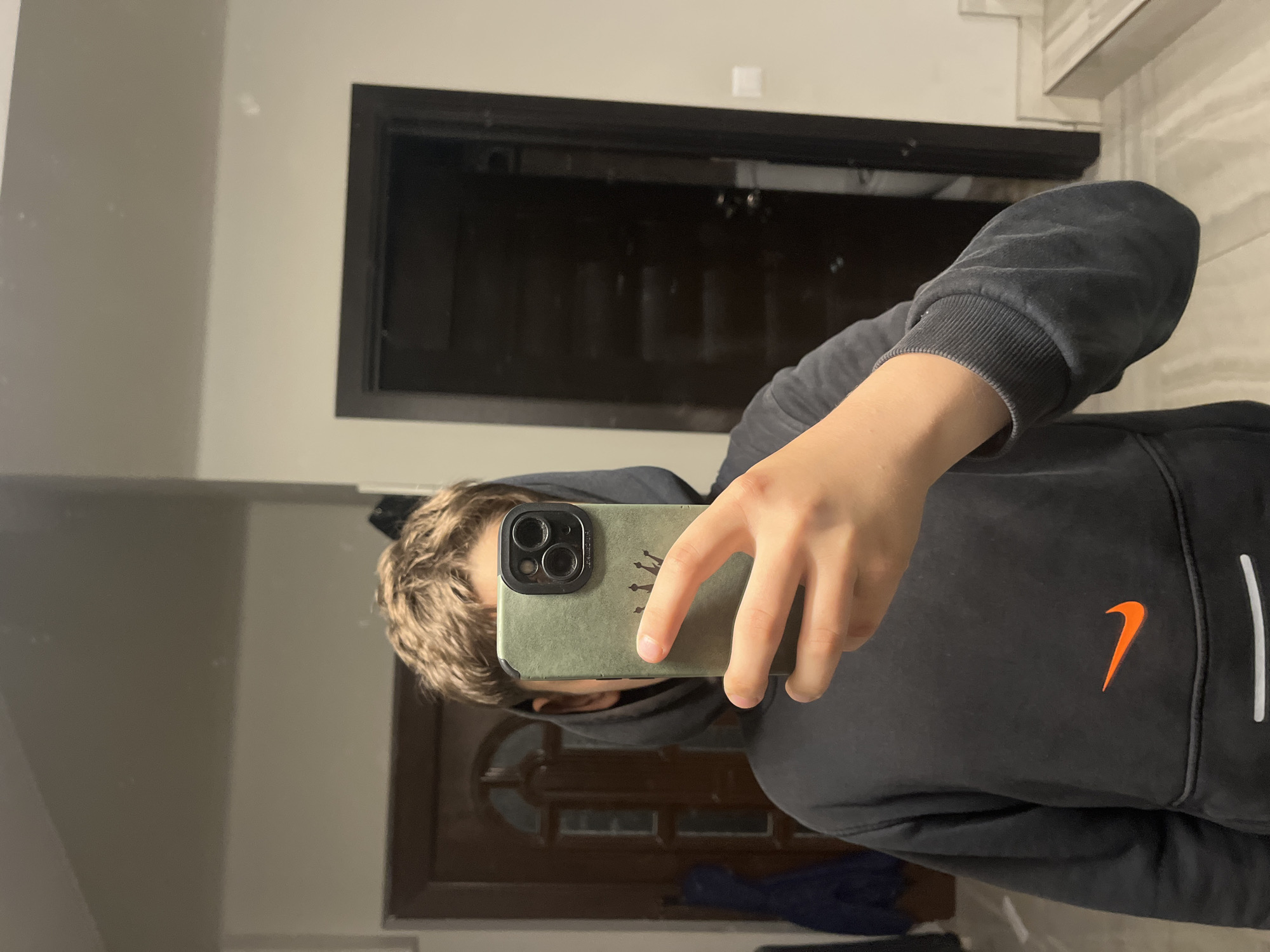 A mirror selfie captures a young individual, likely male, with short, wavy light brown hair, holding a smartphone. The phone is in their right hand and encased in a sage green cover featuring a black triple-lens camera module and a small gold crown logo. The person is wearing a black hoodie, prominent with an orange Nike swoosh logo on the front and a white stripe visible along the side. The ribbed cuff of the hoodie's sleeve is also discernible.
The reflection in the mirror reveals an indoor setting with light-colored, possibly off-white or light gray, walls. Two dark-framed doorways are visible in the background; one is dark and angular, appearing directly behind the person's head, while another, seemingly a wooden door with panelling, is reflected lower down. The lighting suggests an indoor environment, likely artificial. The scene is located in Cherni Vrah, Bulgaria.