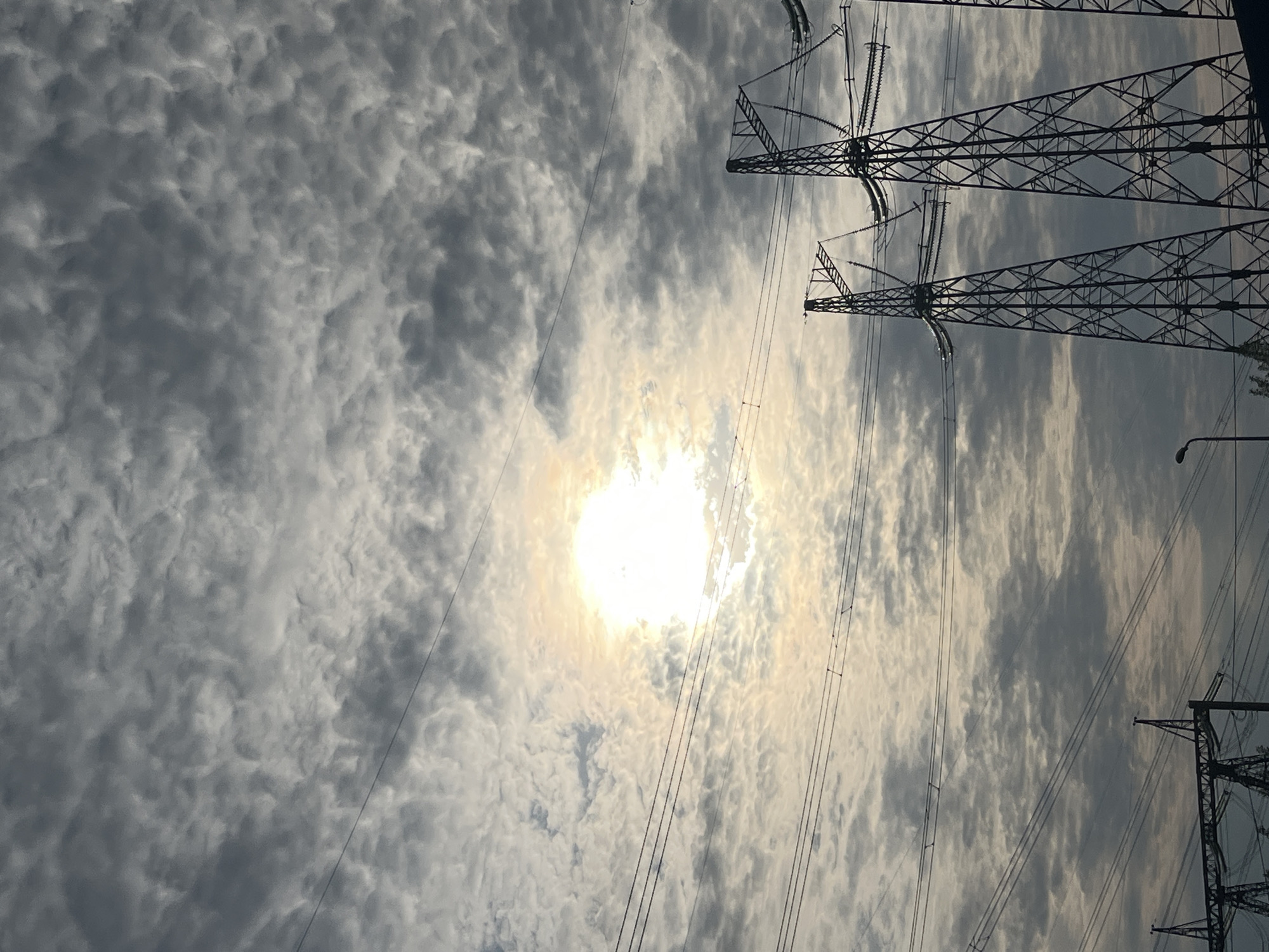 The image shows a cloudy sky with the sun shining through. In the foreground, there are power lines and transmission towers. The location is likely in an industrial or suburban area, possibly near Chaikovsky, Russia, given the prompt's specification.