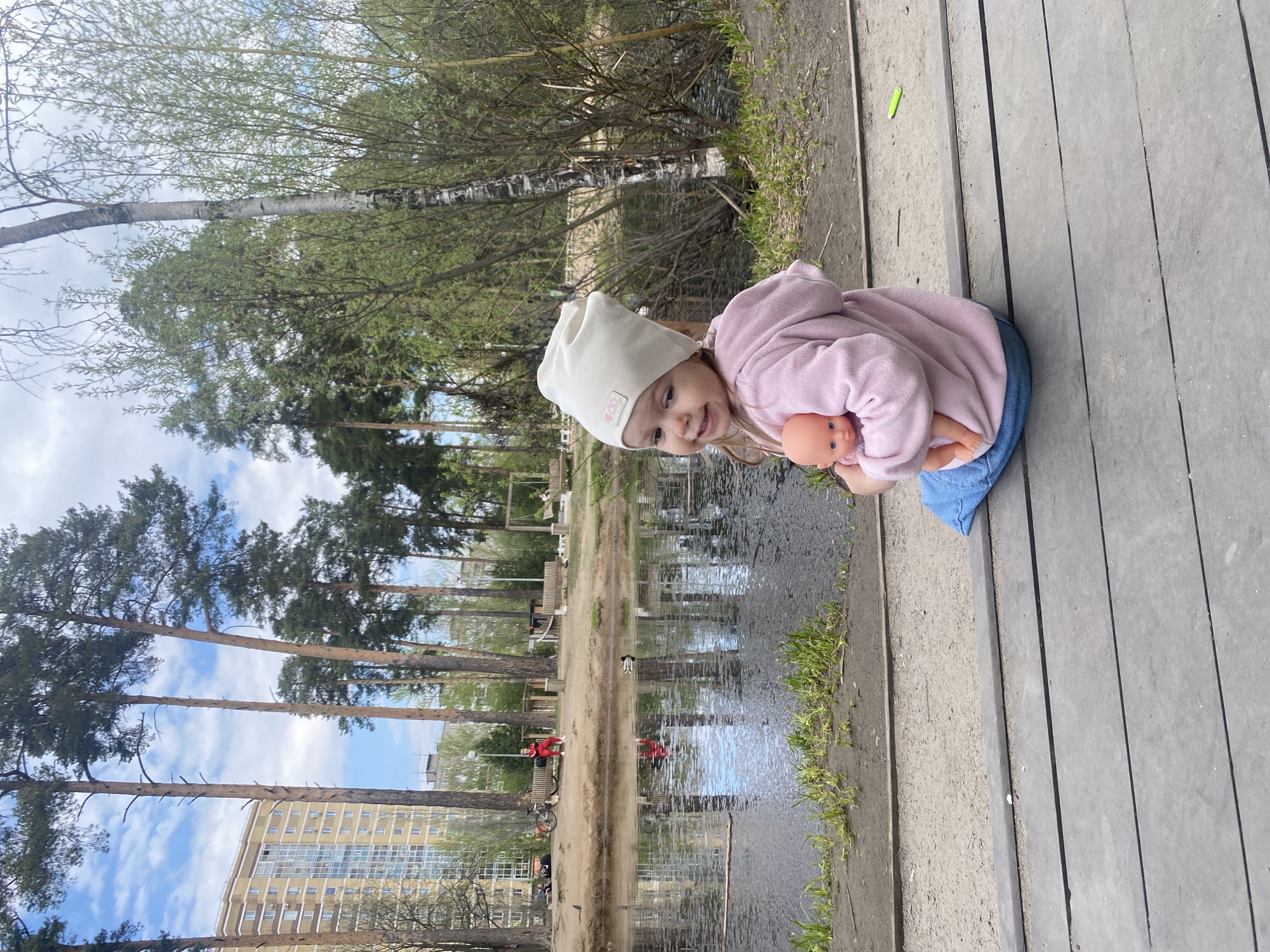 A young girl in a white hat, pink hoodie, and blue pants is seated on a wooden structure next to a body of water. She holds a doll in her hands. Behind her is a view of trees, a building, and reflections in the water. In the background is the city of Perm, Russia.