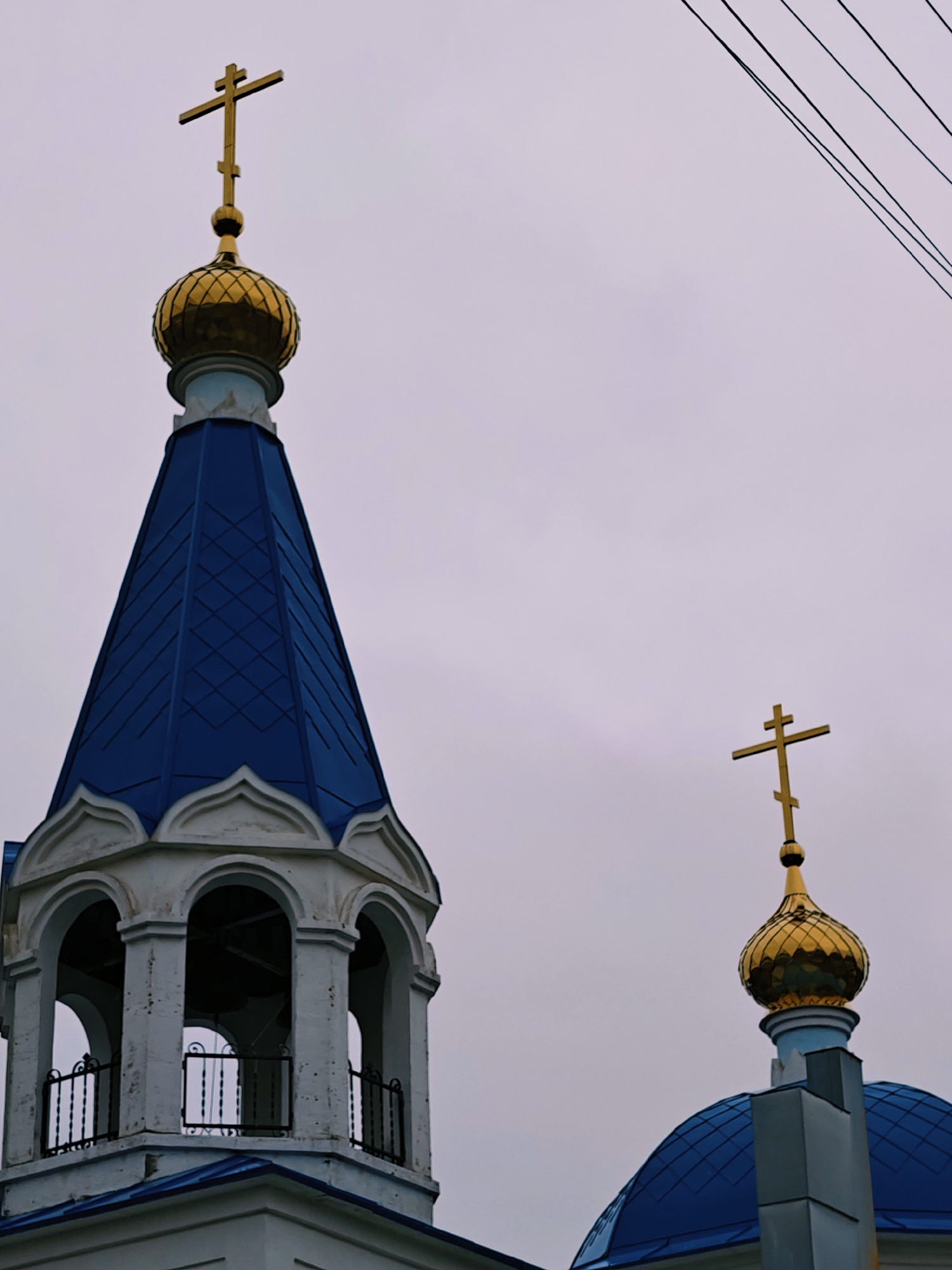 The image displays a partial view of an Eastern Orthodox church building, characterized by its distinctive architecture. Two golden, onion-shaped domes, each topped with an eight-pointed Russian Orthodox cross, dominate the upper portion of the frame against an overcast, light grey sky.
In the foreground and to the left, a prominent bell tower rises, constructed from light-colored, possibly white, stone or stucco. This tower features a section with three visible arched openings, beneath which are dark metal railings, suggesting a belfry or observation level. The tower is capped with a deep blue, conical roof that exhibits a subtle diamond or scale pattern. A larger golden onion dome, surmounted by a golden cross, sits atop this conical roof.
To the right, a smaller, golden onion dome and cross are visible, rising above a blue, hemispherical roof. This structure appears to be part of another section of the church, set further back or lower. The golden surfaces of the domes and crosses reflect the muted light of the cloudy day.
Thin, dark power lines diagonally traverse the upper right corner of the image, contrasting with the pale sky. There are no people visible, and no specific activity or event is depicted. The time of day appears to be daytime, though the sky is uniformly cloudy, indicating overcast weather. No text is visible in the image. The architectural style points to a location such as Noviy, Russia.