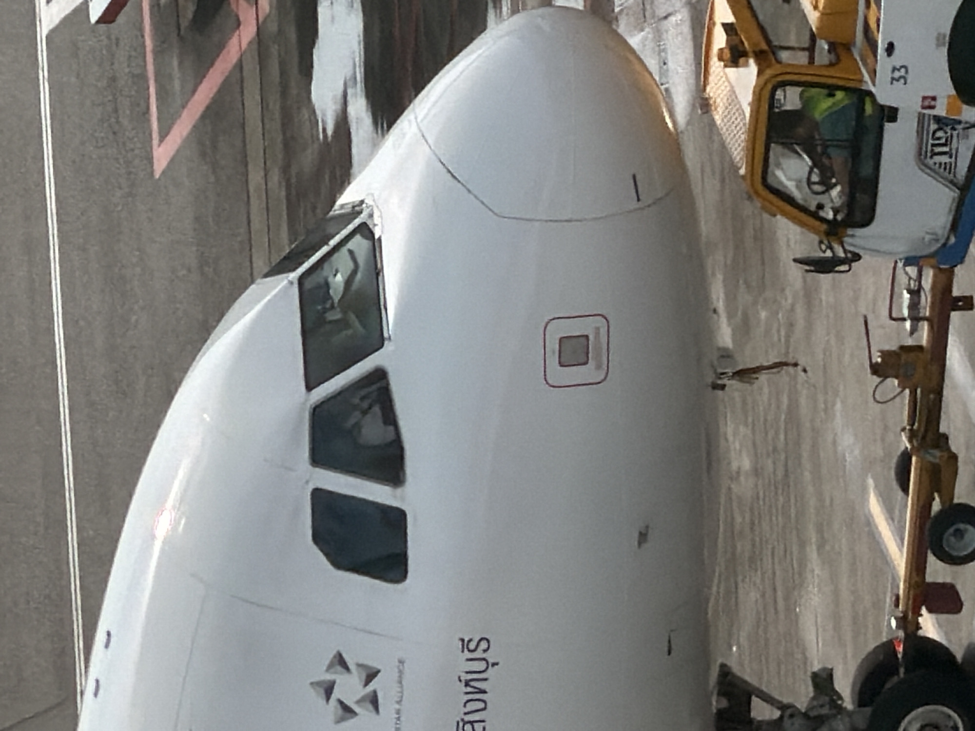 An airplane is parked on the tarmac. The aircraft is white with the Star Alliance logo and text in Thai script. The cockpit windows are visible, and a person is seated in the pilot's seat. A tow truck is connected to the front landing gear of the airplane. The tow truck is yellow and white and has the number "33" printed on its side. The tarmac is gray and wet, with red lines painted on it. The overall scene is indicative of an airport environment.