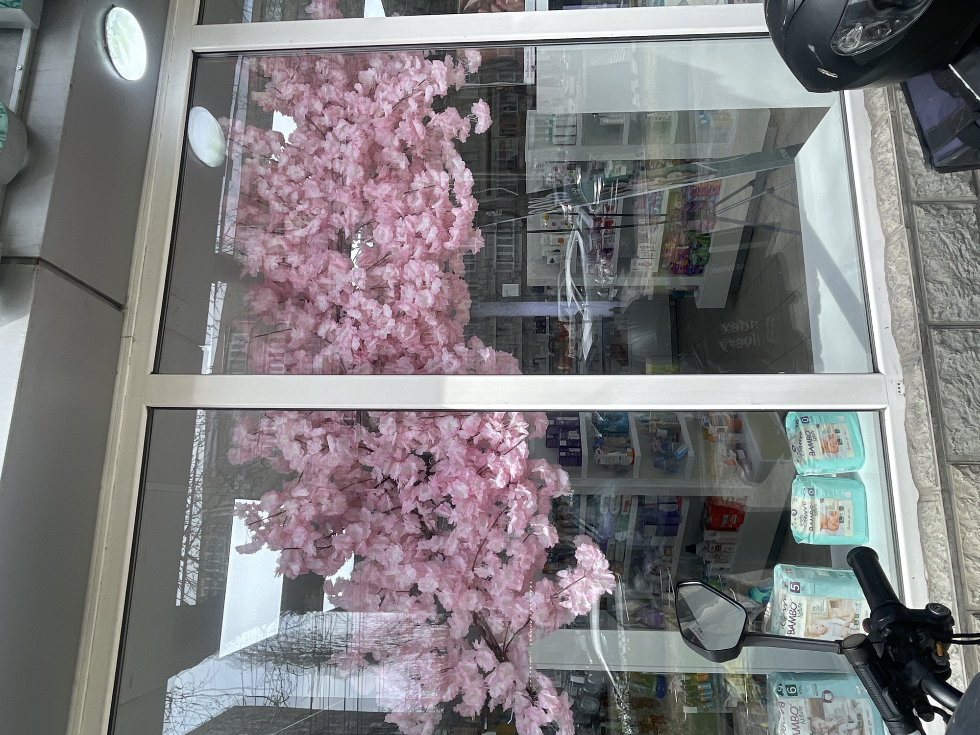 The image captures the exterior of a business establishment, likely a pharmacy, as suggested by the shelves stocked with various products visible through the glass windows. A decorative pink cherry blossom tree is prominently displayed within the establishment, and its reflection is visible on the glass surface. A motorcycle is parked outside the building, with its black helmet, handlebars, and side mirror partially obscuring the view. The location appears to be Yerevan, Armenia, as inferred from the Armenian text visible on one of the product packages ("BAMBOO Nature") displayed on the shelves. The building has a light-colored facade, and the street outside is paved with light gray tiles. The scene suggests a commercial area during daylight hours.