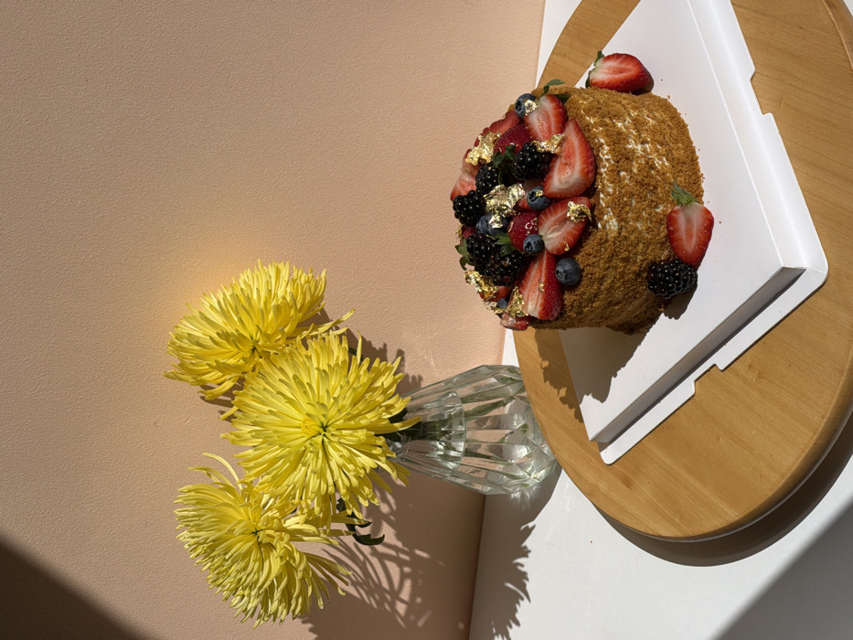 The image depicts an indoor still life composition. On the left, a clear, faceted glass vase contains three large, vibrant yellow spider chrysanthemums. The vase rests on a white horizontal surface, casting a distinct shadow to its left. On the right, a small, round, multi-layered cake, approximately 4-5 inches tall, features light brown, crumb-coated sides. The top of the cake is generously adorned with fresh berries, including sliced strawberries, whole blueberries, and whole blackberries, interspersed with small flakes of edible gold leaf. A whole strawberry, a sliced strawberry, and a whole blackberry are also present on the white square cardboard cake board supporting the cake. This cake board rests on a larger, light-colored round wooden serving tray. The background consists of a plain, textured peach-colored wall. Strong, directional lighting from the upper left illuminates the scene, creating prominent, sharp shadows on the white surface and a diagonal shadow across the background wall. No individuals are present, and no actions or interactions are depicted. The specific location within Tel-Aviv, Israel, is not discernible from the visual information provided.