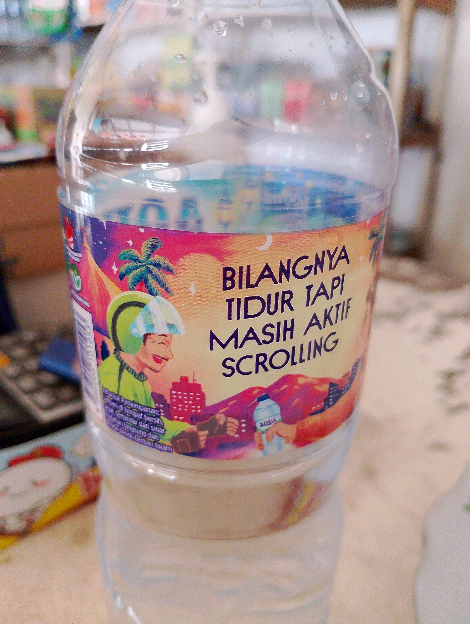 A clear plastic bottle of Aqua mineral water sits on a table, its label featuring a colorful cartoon illustration. The illustration depicts a man in a green helmet and jacket, riding a motorcycle. Behind him, a stylized cityscape with buildings and palm trees is visible, set against a gradient sky of pink, orange, and blue. Text on the label reads, "BILANGNYA TIDUR TAPI MASIH AKTIF SCROLLING," which translates to "Says sleeping but still actively scrolling." The bottle appears to be partially full. The background is blurred, showing hints of shelves with colorful items and a keyboard. There is no identifiable location in Merauke, Indonesia, visible in the image.