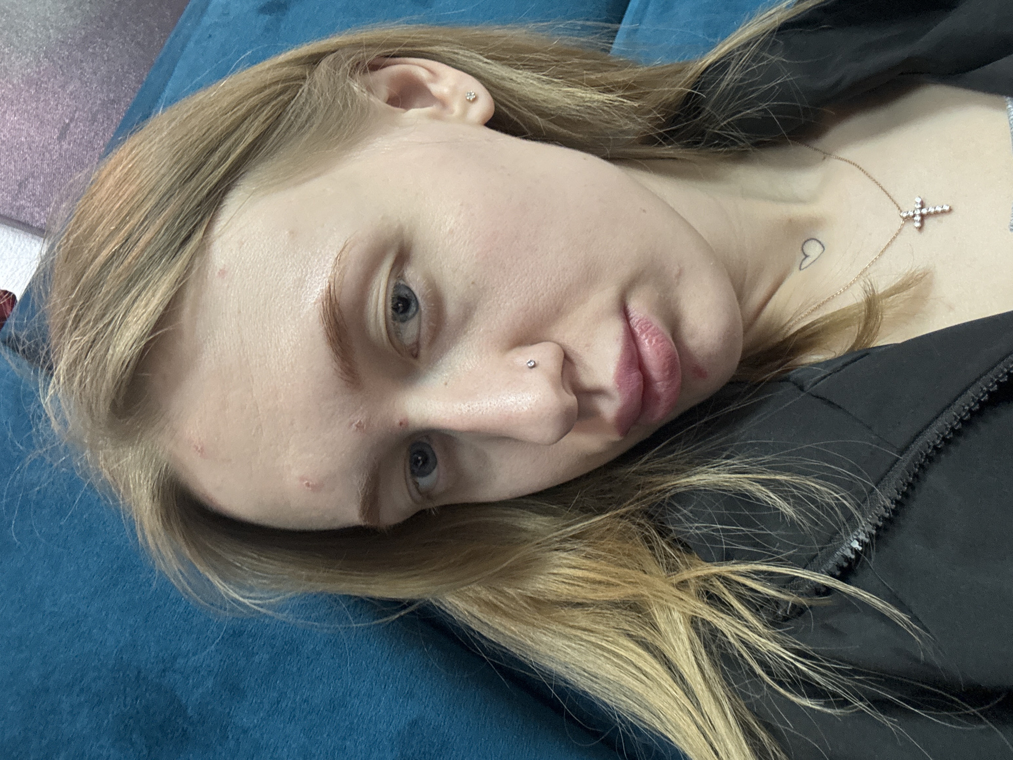 A fair-skinned woman with blonde hair lies on a blue surface, possibly a couch or bed. She is wearing a black jacket or top. The woman has light blue eyes, and her face shows some blemishes. She has a nose piercing and earrings. A gold necklace with a cross pendant is visible on her chest. A small heart tattoo is located near her collarbone. The location is not discernible in the image.