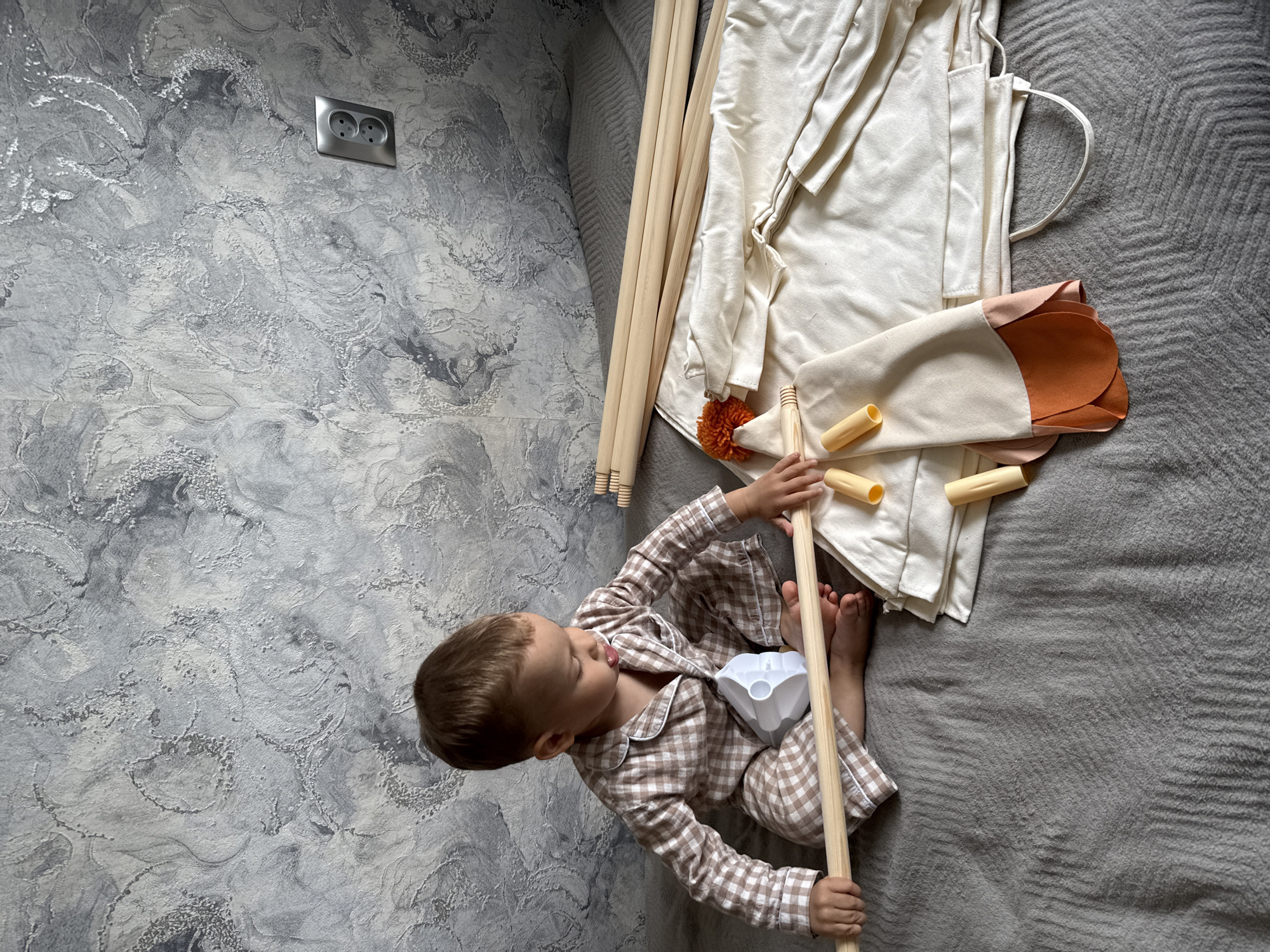 A high-angle shot depicts a young child assembling a play tent on a gray textured surface that appears to be a bed or mattress. The child, wearing brown and white plaid pajamas, sits amidst the tent's components, including several wooden poles and a white canvas tent cover. A small, white plastic piece is also present, likely part of the tent's assembly. The background features a gray, textured wall with a double electrical outlet. This scene is located in Шебунино, Russia.