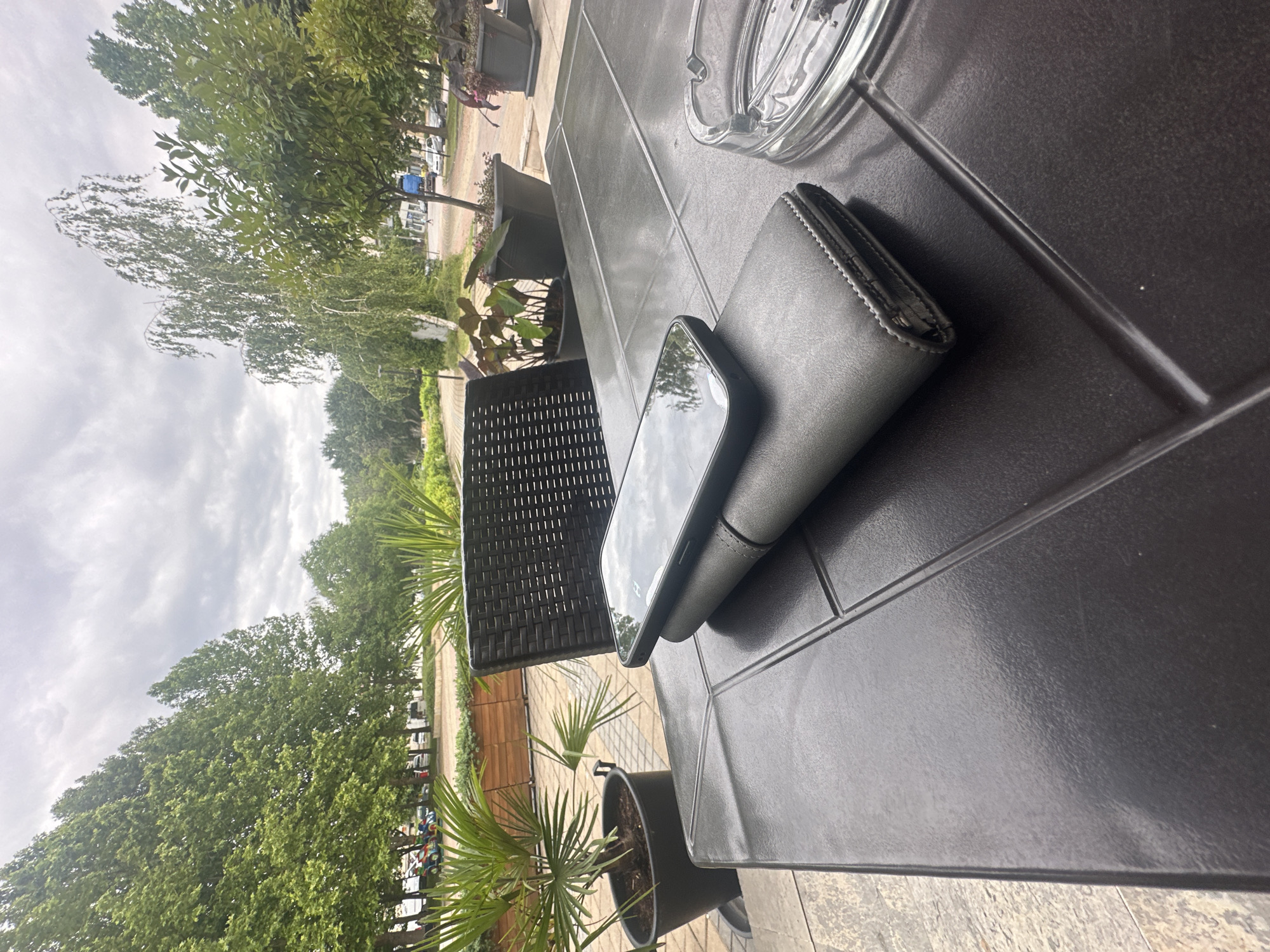 The image captures an outdoor scene from a low-angle perspective, looking outwards from a table. In the immediate foreground, a dark-colored table, possibly made of plastic or composite material, occupies the right half of the frame. Resting on this table is a black smartphone, partially obscuring a dark grey wallet or phone case beneath it. To the upper right, a clear glass ashtray is visible.
To the left, a section of a black wicker chair can be seen, hinting at a seating arrangement. Several potted plants, including small palms and other leafy varieties, are positioned around the table area, contributing to a relaxed outdoor ambiance.
The background unfolds under an overcast sky with scattered clouds, indicating daytime. A long line of dense, green deciduous trees stretches across the midground, bordering what appears to be a road or pathway. Several parked cars are partially visible along this thoroughfare, suggesting a public or accessible location.
The setting appears to be an outdoor seating area, possibly a patio of a cafe or restaurant, in Lom, Bulgaria. The objects on the table suggest someone was momentarily seated there. No people are visible, and no discernible text appears on any object. The overall impression is one of a calm, cloudy day at an outdoor venue.