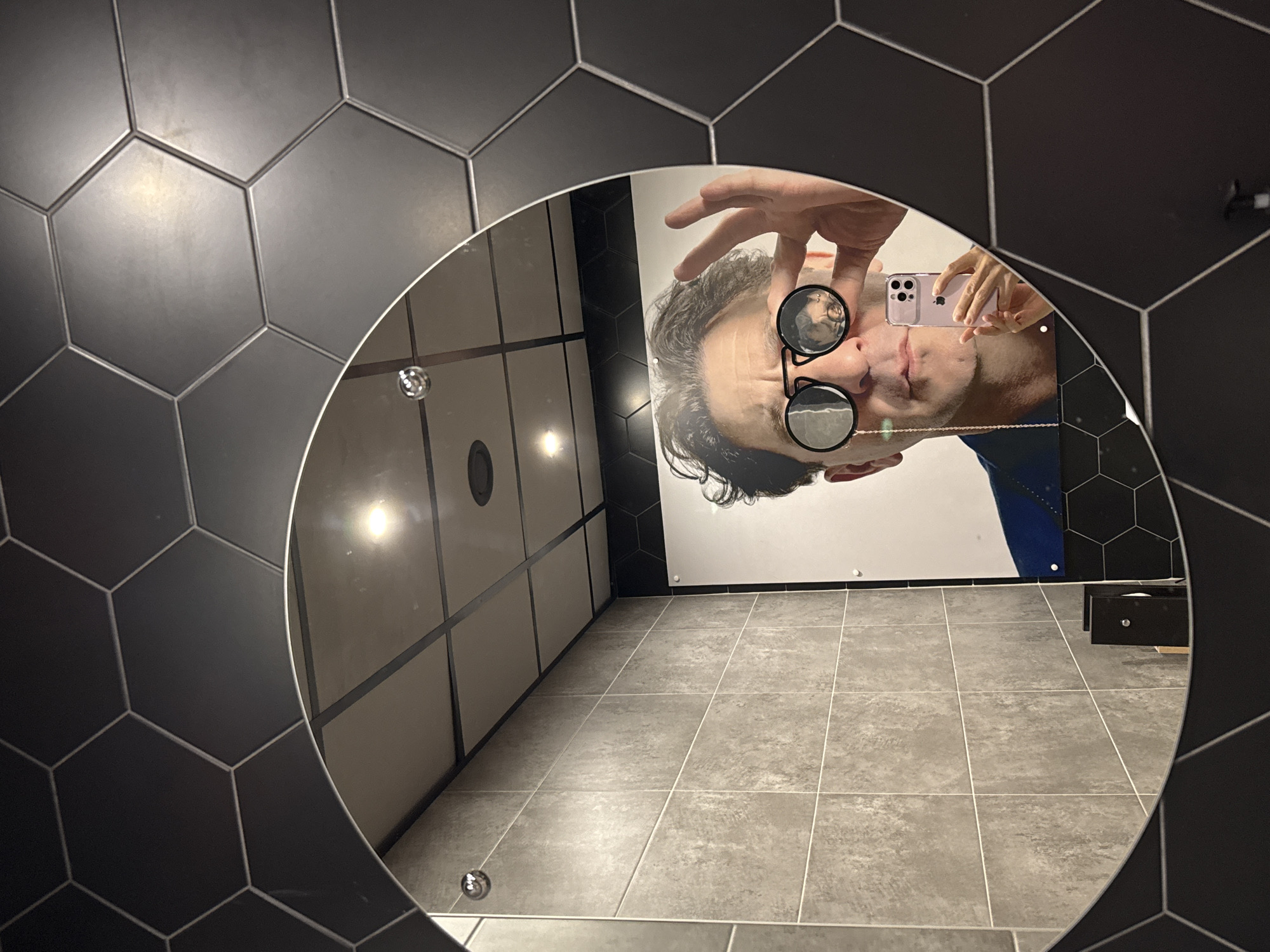 This is a mirror selfie taken in a public restroom. The subject, a man wearing round, black-rimmed glasses and a blue jacket, is holding a light purple smartphone to take the photo. He is also holding one of his hands up to adjust his glasses. A chain hangs from his glasses to his collar. The mirror is circular and framed by hexagonal black tiles. The background of the restroom includes grey tiled walls and a tiled ceiling with lights. A large poster of the man is visible on the wall behind him. The location is not specified as Kirishi, Russia; this information is not present in the image.