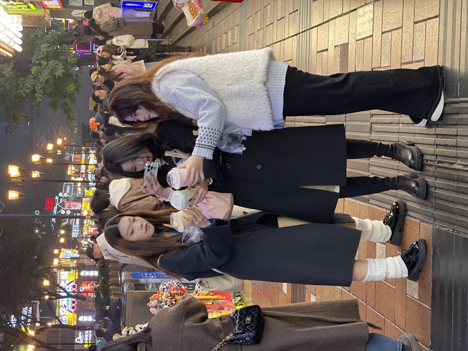The image displays a nighttime street scene within Yuzhong District, China, illuminated by numerous streetlights and vibrant commercial neon signs. Three young women occupy the foreground on a paved pedestrian walkway.
The woman on the left, with long dark hair, wears a dark long coat, white leg warmers, and black patent leather platform shoes. She holds two clear plastic cups containing light-colored beverages with straws, and a light-colored shoulder bag is visible.
The central woman, also with long dark hair, wears a dark long coat and black platform boots. She holds a white smartphone horizontally with both hands, positioned as if taking a photograph of the beverages, and simultaneously holds one clear plastic cup with a straw.
The woman on the right, possessing long reddish-brown hair, wears a light grey sweater with patterned cuffs, a white fluffy vest, black pants, and black and white platform sneakers. She holds one clear plastic cup with a straw.
The women are clustered around the beverages, with the central woman's action focused on photography.
The background reveals a densely populated urban environment with numerous pedestrians. Buildings lining the street display brightly illuminated signs featuring Chinese characters. Ornate streetlights emit a warm orange glow, and scattered trees with sparse foliage are visible. On the far left, a vendor's stall exhibits colorful items, and a person wearing a brown coat and a black quilted shoulder bag with a gold chain strap is partially visible. The ground is paved with light-colored rectangular tiles and includes textured tactile paving strips.