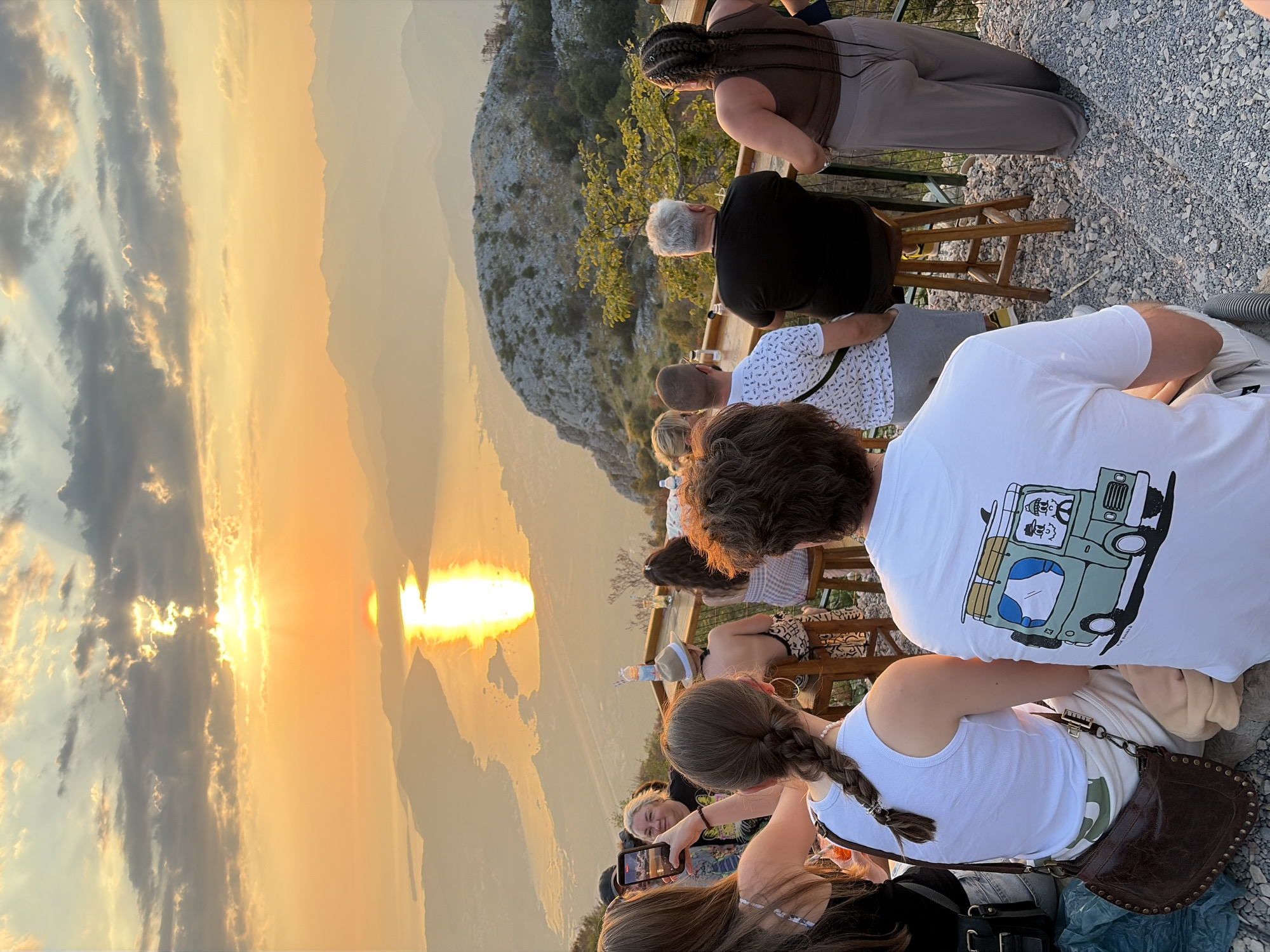 In Kuk, Montenegro, a group of people is gathered at a scenic overlook, likely a restaurant or bar, to watch the sunset. The sun, a bright golden orb, is low in the sky, casting a brilliant reflection on the water below and illuminating the mountainous landscape. The sky is filled with dramatic clouds, streaked with orange and yellow hues.
Several individuals are seated at a high counter, their backs to the camera, facing the panoramic view. One person, wearing a black t-shirt, is seated on a wooden stool. To their right, a woman with braided hair is standing, wearing a brown tank top and grey pants. Another woman, also with braided hair, is seated in the foreground, wearing a white tank top and cream-colored pants. She has a brown leather handbag with studded details resting beside her. The foreground is composed of gravel and stones, with some foliage visible.
In the middle ground, more people are seated at the counter, some holding drinks. One woman is taking a photo of the sunset with her smartphone. A man in a white t-shirt with a printed pattern is also visible. The overall atmosphere suggests a relaxed and social gathering, appreciating the natural beauty of the location. The distant mountains are layered and hazy, fading into the atmospheric perspective of the sunset.