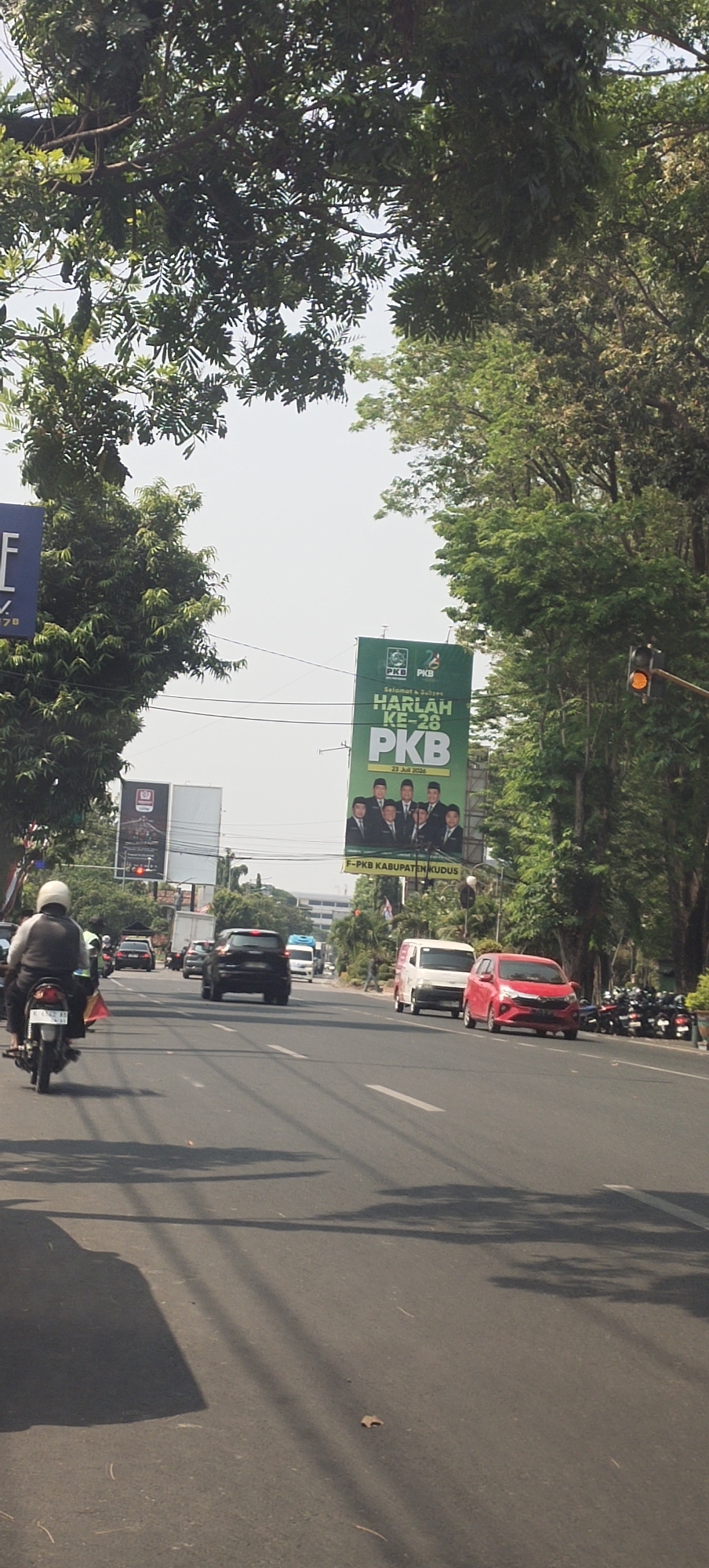 The image captures a street scene in Jati Kulon, Indonesia, dominated by a wide road with multiple lanes, lined by mature trees on either side. The sky is bright and overcast, suggesting daytime.
In the foreground, a motorcycle rider, wearing a white helmet and a dark jacket, is moving away from the camera in the left lane. The motorcycle's license plate is partially visible.
Further down the road, a line of vehicles is visible. In the right lane, a red car is prominent, followed by a white van and then a black SUV. A few other cars and a white truck are also present in the middle and left lanes. On the right side of the road, a group of parked motorcycles is lined up.
Dominating the right side of the frame, a large green billboard is erected above the trees. It features a political advertisement with the text "SELAMAT & SUKSES HARLAH KE-26 PKB" and "F-PKB KABUPATEN KUDUS" along with portraits of several men in suits. A traffic light with an orange signal is visible to the right of the billboard. On the left side of the road, another billboard, partially obscured by trees, displays a blue background with white text.
The road surface is asphalt, with white lane markings clearly visible. Shadows from the trees are cast across the road. The environment appears to be a semi-urban or suburban area, indicated by the presence of traffic, billboards, and residential or commercial buildings in the background.