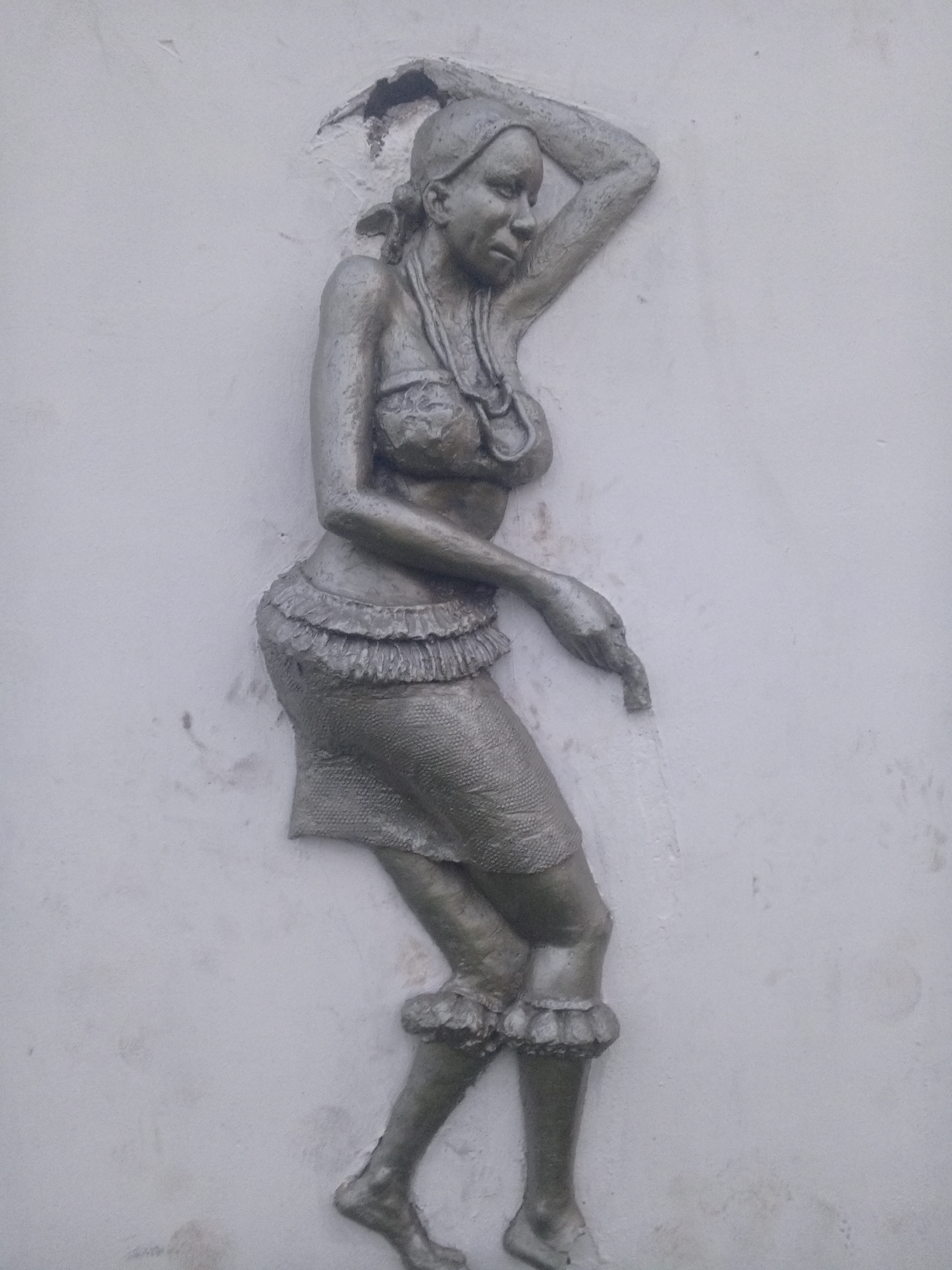 A bas-relief sculpture of a woman is mounted on a white wall. The woman is depicted in a dance pose with her right arm raised above her head and her left arm extended downward. She wears a textured skirt with a ruffled waistband and ruffled anklets. Her breasts are covered with a similar textured fabric. A beaded necklace adorns her neck, and her hair is pulled back. The sculpture is silver or gray in color, contrasting with the white background. The precise location in the city of Uyo, Nigeria is not discernable from the image provided.