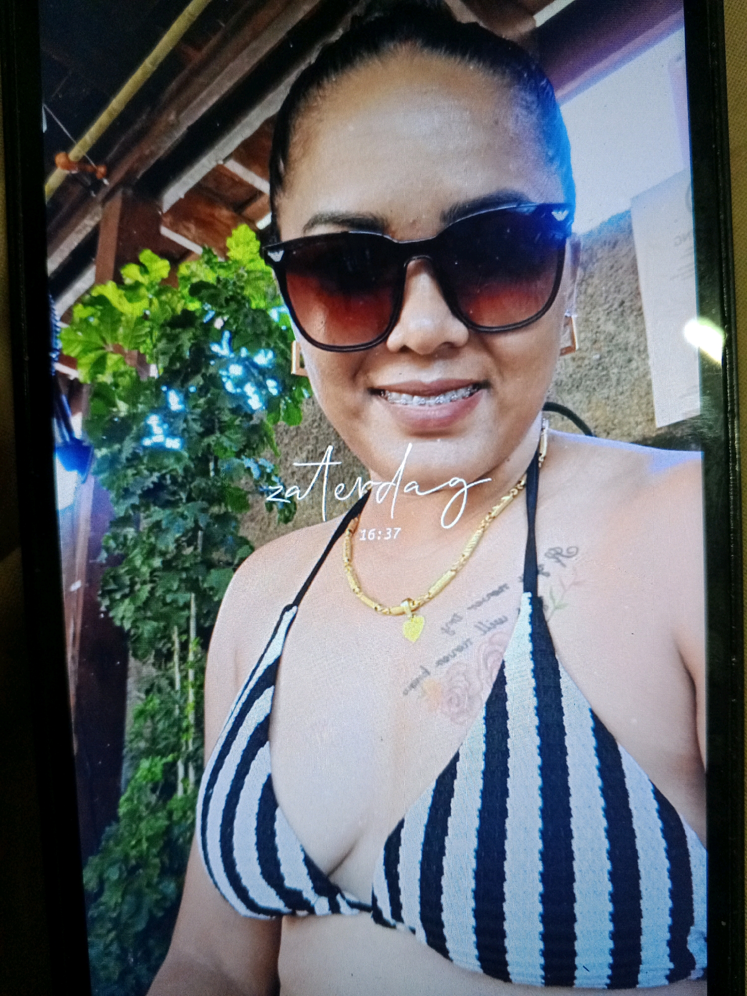 A close-up shot features a woman wearing sunglasses and a striped bikini top, with braces on her teeth. She is adorned with a gold necklace and earrings. Tattoos are visible on her shoulder and chest. A digital clock on the screen displays "16:37" and the word "zaterdag" (Dutch for Saturday). The background reveals lush green foliage and a wooden structure, suggesting an outdoor setting. The woman is positioned in the foreground, occupying most of the frame. The overall scene appears to be a candid photograph taken in Noord, Suriname, possibly at a resort or residential area given the tropical vegetation.