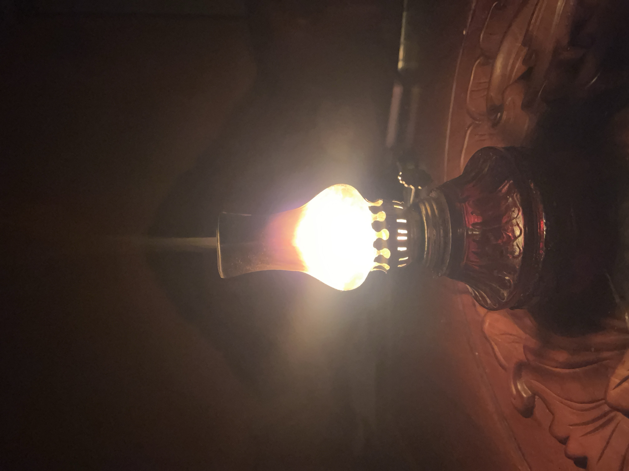 The image shows a lit oil lamp. The lamp is composed of a red glass base and a white, bulbous glass chimney, with a metal burner assembly in between. The lamp is sitting on a dark, possibly wooden surface with intricate carvings. The background is dark, obscuring any further details about the location. Based on the prompt, this scene is located in Lhokseumawe, Indonesia. However, the image itself provides no specific evidence to confirm this geographic location.