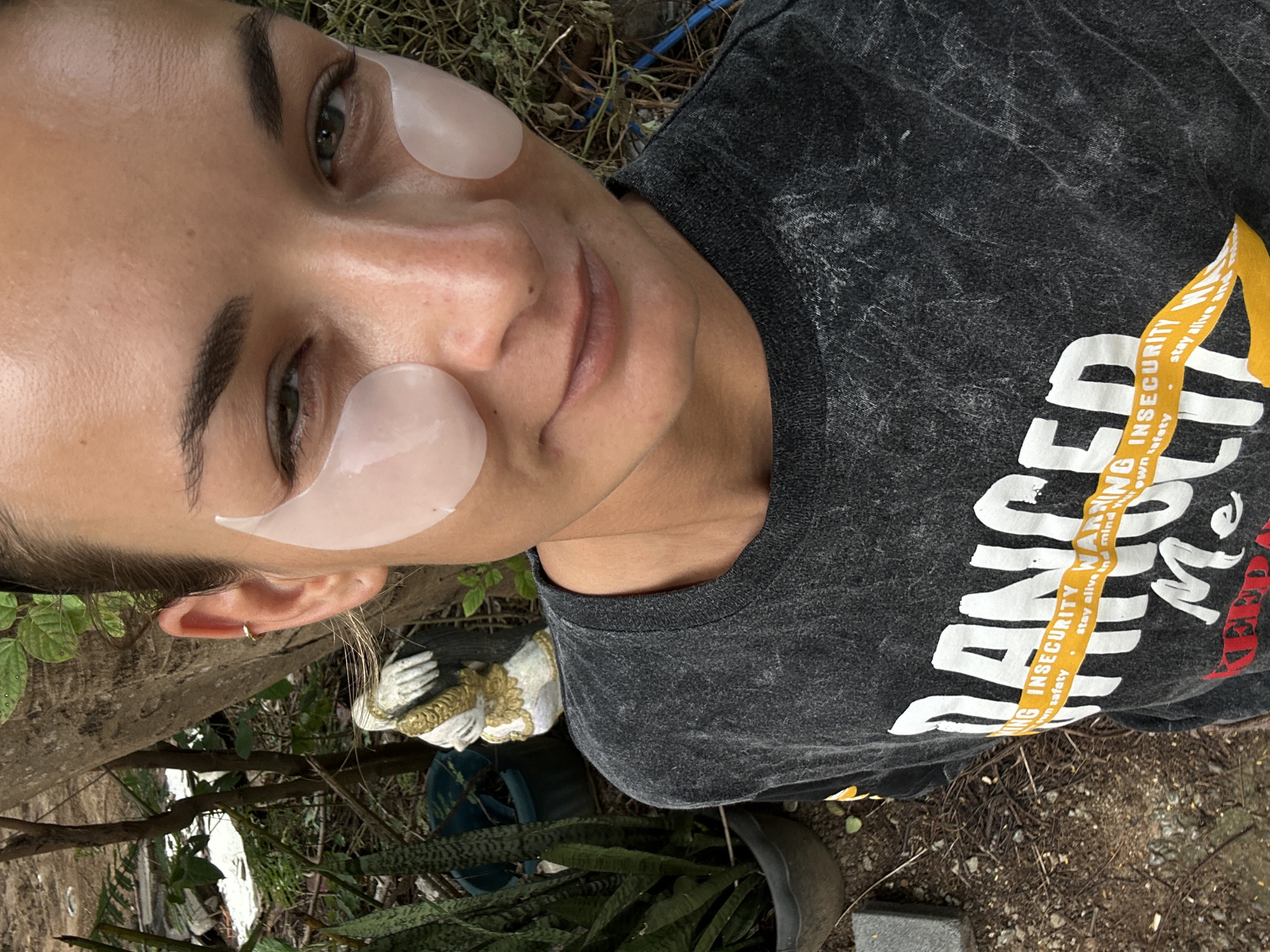 The media file shows a close-up, vertically oriented, selfie-style shot of a woman's face and upper chest. She has dark eyebrows and is wearing light-colored under-eye patches. Her eyes are open and she is looking towards the camera with a slight smile. She is wearing a dark gray, distressed t-shirt with a graphic design. The text visible on the t-shirt reads, "DANCEO," "ING INSECURITY WARNING," and "stay alive and mind Your own safety." The background appears to be an outdoor setting with foliage, branches, and what looks like a small garden statue. The lighting suggests it could be daytime. The location is identified as Kathu, Thailand.