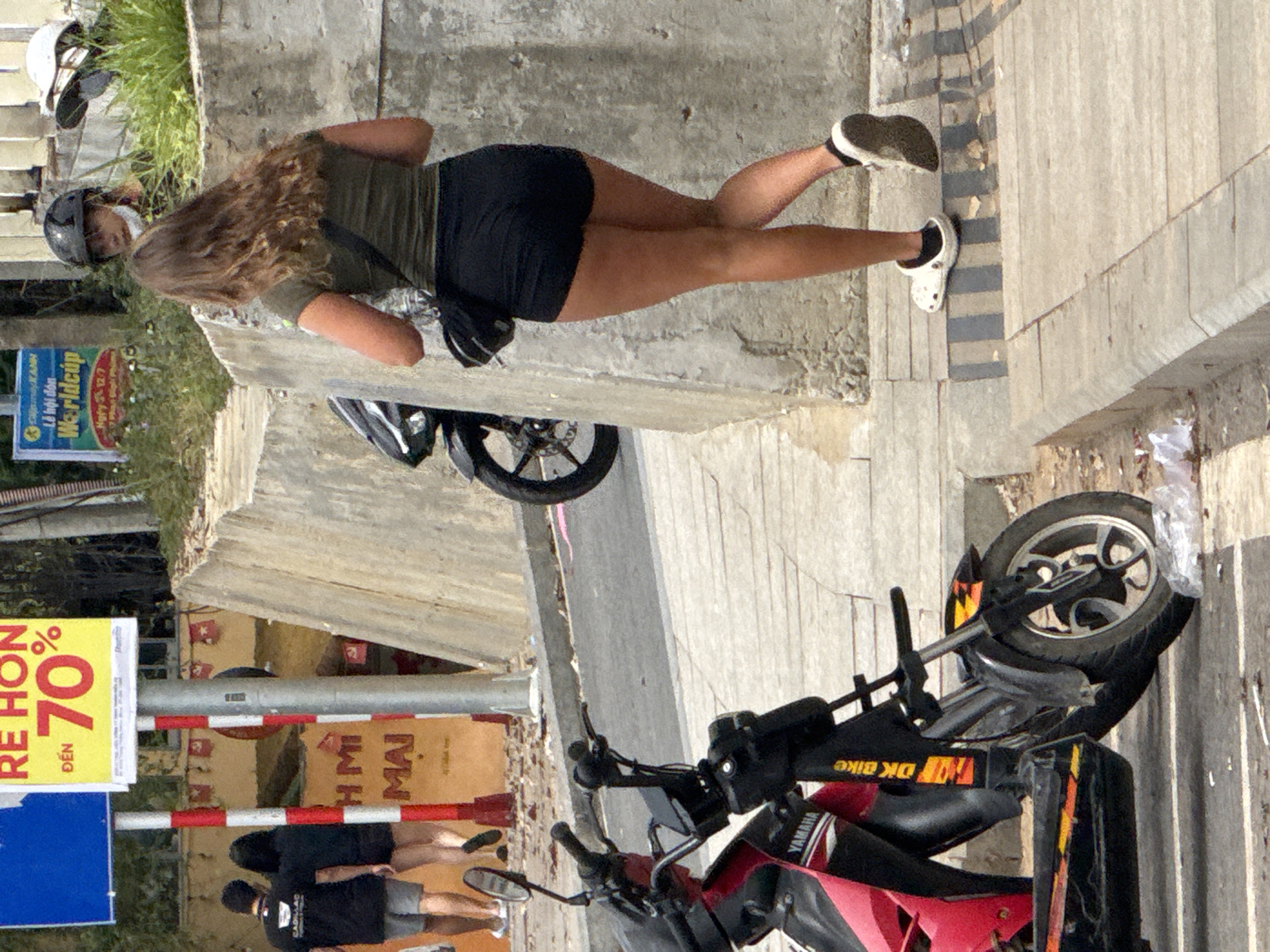 The image depicts a street scene in Da Lat, Vietnam, focusing on a woman walking away from the camera. She has long, wavy brown hair and is wearing a dark green t-shirt and black shorts. A black crossbody bag is slung over her shoulder. She is wearing white Crocs on her feet. In the background, two individuals, a man and a woman, are walking away from the camera, also wearing dark clothing. A motorcycle is parked on the left side of the image, with "YAMAHA" visible on its side. To the right, a man wearing a helmet and a white medical mask is partially visible. A sign with Vietnamese text, including "RE HON DEN 70%", is prominently displayed on the left, indicating a sale. The environment appears to be a sidewalk adjacent to a street, with concrete barriers and some vegetation visible.