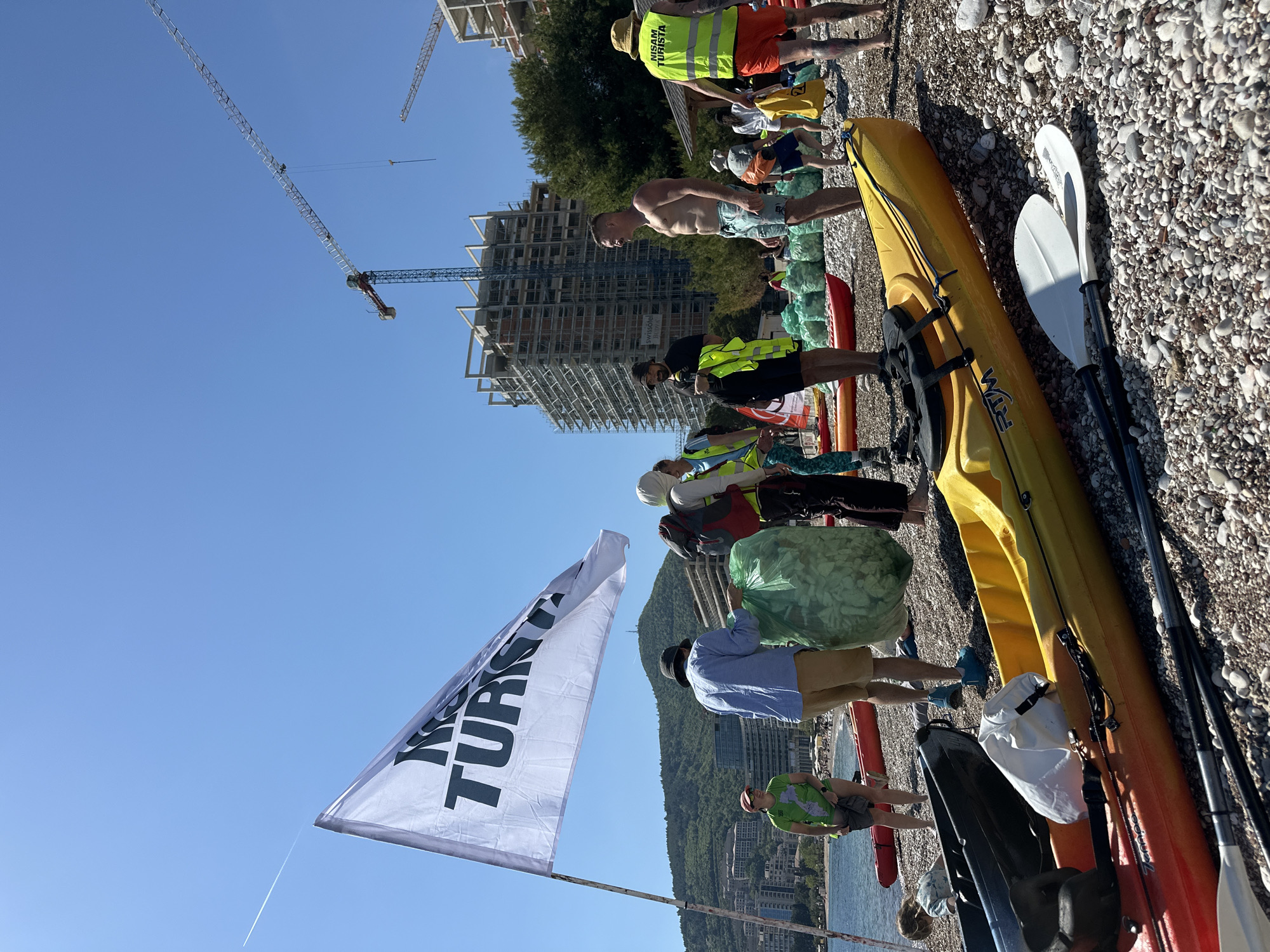 A group of people are on a pebble beach in Budva, Montenegro, with kayaks and trash bags. The group appears to be collecting trash. A flag with "I TURISTI" is visible. In the background, there is a construction site with multiple buildings under construction and several cranes. The sky is clear and blue.