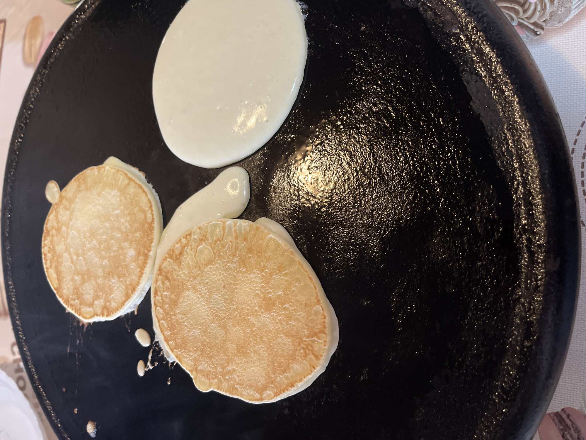 The image shows a black, presumably non-stick, crepe pan or griddle. There are three portions of pancake batter on the pan. Two of the portions have been cooked to a light golden-brown on at least one side. One of the pancake portions is uncooked and white in color. A small stream of batter has flowed from the uncooked batter to the partially cooked pancake. The image does not include sufficient details to conclusively determine the location.