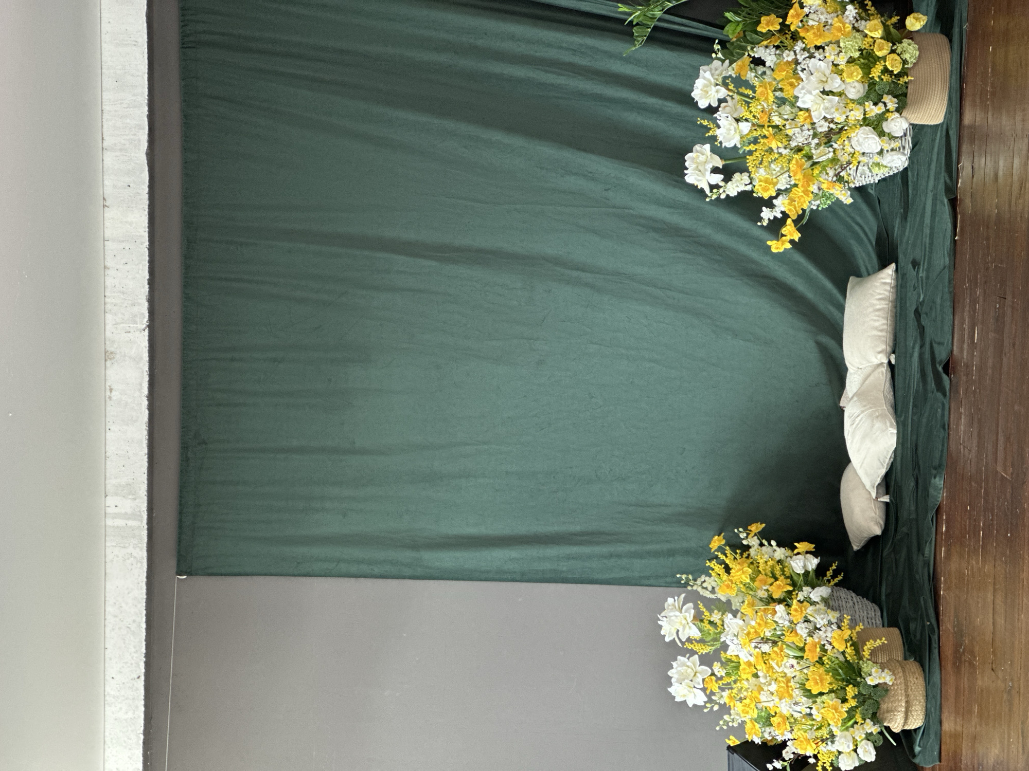 The image shows a staged indoor setting. A backdrop of draped dark green fabric dominates the scene, occupying the upper portion of the image. The lower portion features a gray wall. Baskets filled with yellow and white flowers are positioned in the upper-right and lower-right corners of the image. To the right of the green fabric are some pillows and the edge of a wooden floor. The image appears to be taken in a studio or controlled environment for photographic purposes. The exact location in Chaikovsky, Russia is indeterminable.