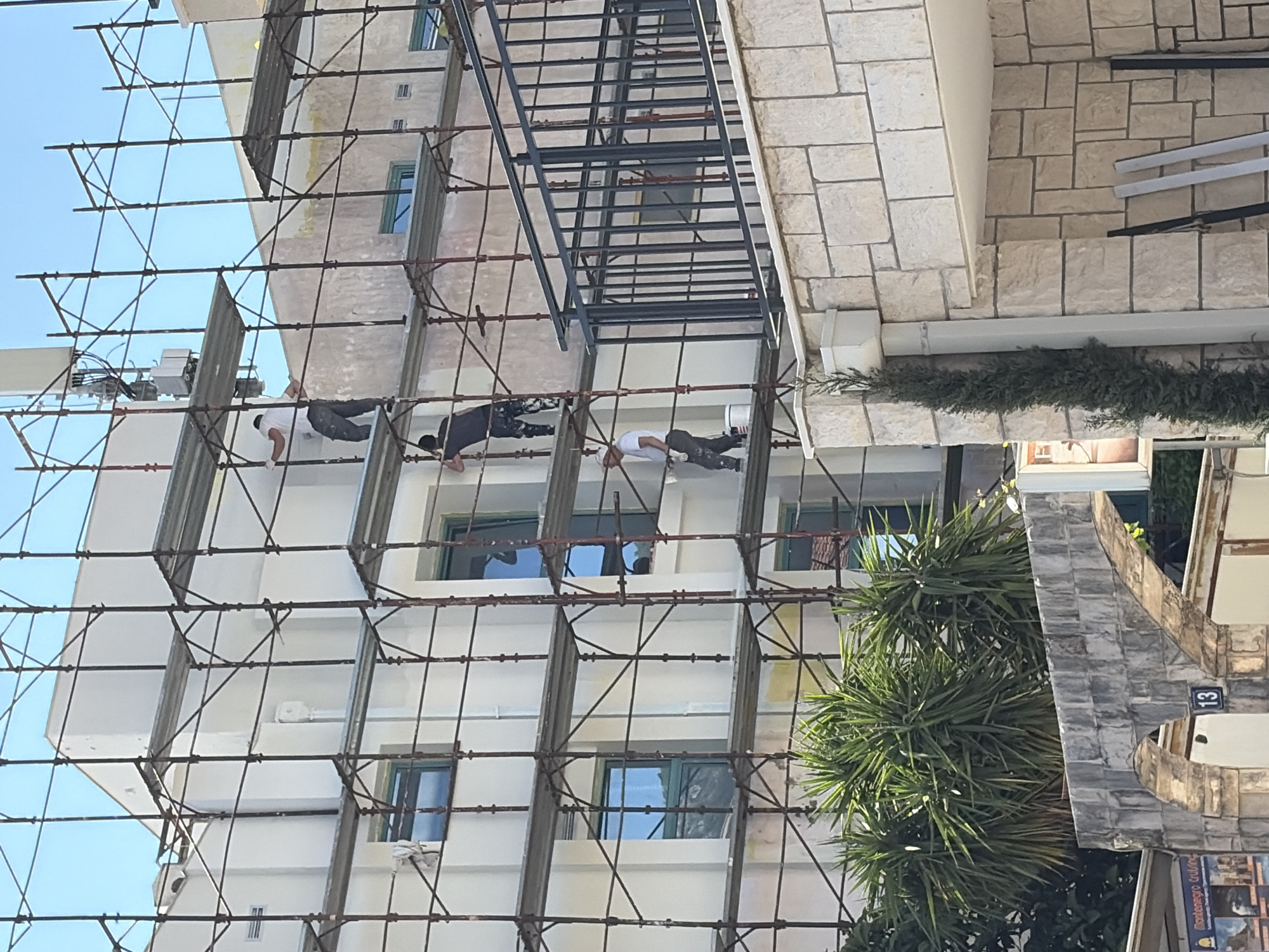 The image shows a multi-story building under renovation in Budva, Montenegro. Three construction workers are working on scaffolding erected along the building's facade. The building is predominantly white with blue-framed windows. Adjacent to the building are green palm trees and a stone structure with the number "13" visible on a plaque. The sky is clear and blue.