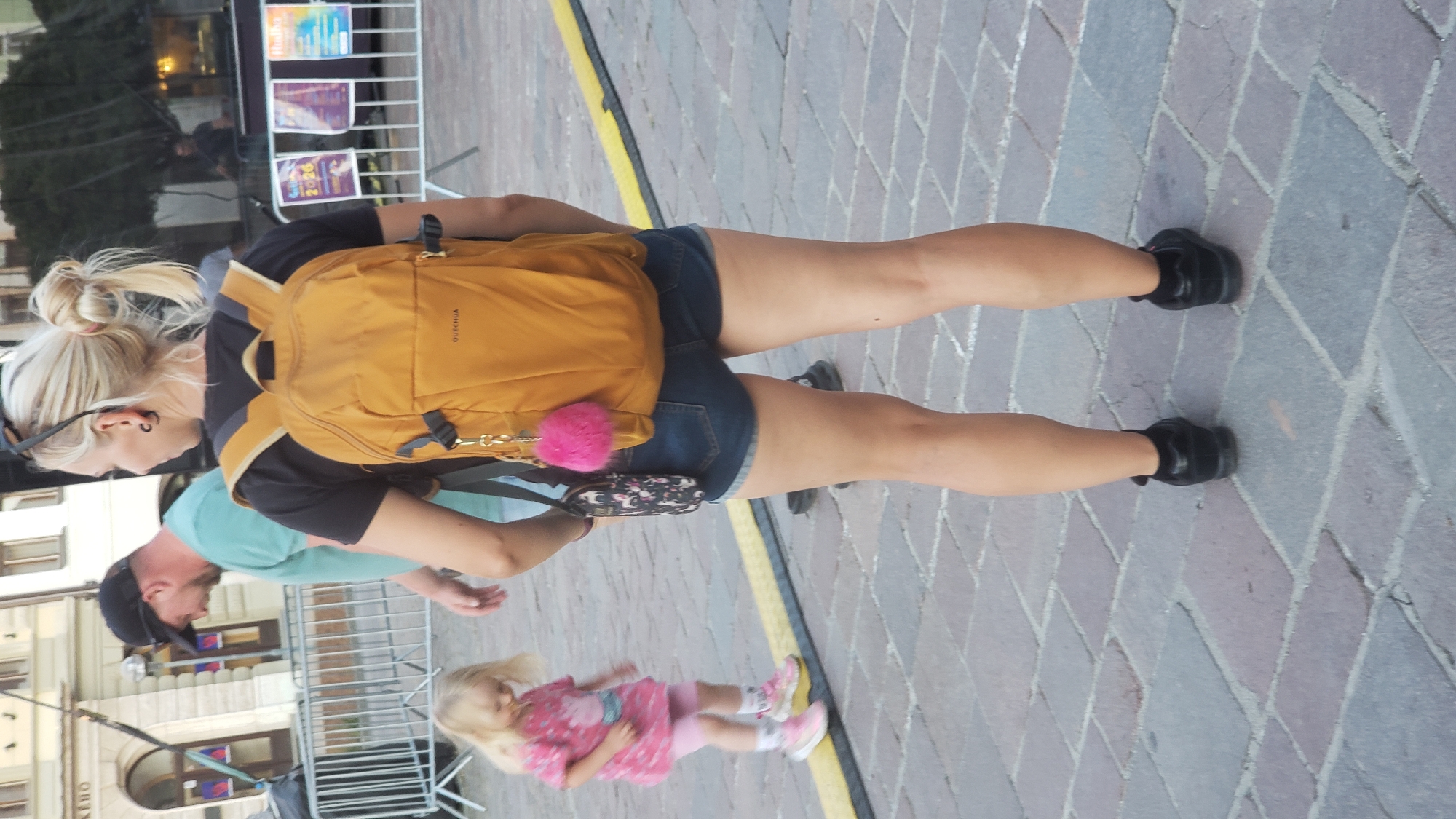 A woman with blonde hair pulled into a bun, wearing a black t-shirt, blue denim shorts, and black shoes, stands on a cobblestone street. She has a yellow backpack slung over her shoulder and is wearing dark sunglasses on her head. A small pink pom-pom charm hangs from her backpack.
Beside her, a man wearing a light blue t-shirt and a black baseball cap is visible, looking down. In the foreground, a young blonde girl in a pink dress and pink shoes is walking.
Behind the woman, a metal barrier separates the cobblestone path from a building facade with several windows. There are posters displayed on a rack in the background.
The scene appears to be outdoors, likely during the day, given the lighting. The cobblestone street and building architecture suggest an urban European setting. The overall atmosphere seems casual, possibly a public space where people are walking around. There is no visible text in the image.
