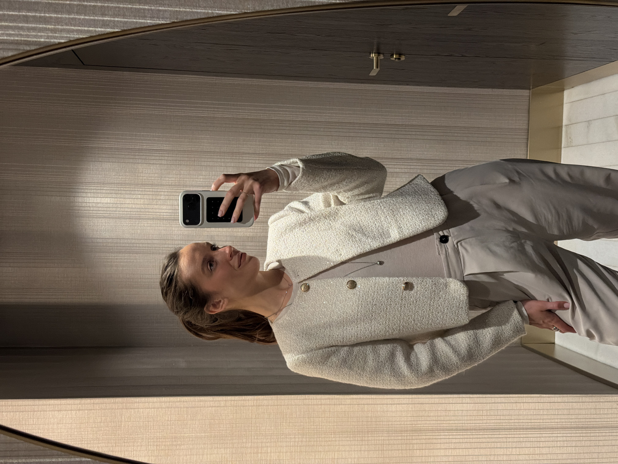 A young woman is depicted taking a mirror selfie indoors, likely within a modern hotel or upscale residence in Budva, Montenegro. She is shown from the chest upwards, holding a light-colored smartphone with a prominent camera array in her right hand, directed towards her reflection. Her left hand is casually tucked into the pocket of her light gray or taupe wide-leg trousers.
She is dressed in a chic ensemble featuring a textured, off-white or cream-colored jacket adorned with gold-toned buttons, worn over a plain, lighter-hued long-sleeved top. A subtle necklace with a small pendant rests around her neck. Her brown hair is neatly pulled back, and her gaze is directed towards the phone, capturing a neutral, composed expression.
The interior setting is characterized by a horizontally striped, light beige or cream wall covering visible behind her in the reflection. Above her, the mirror reflects a dark, possibly wood-paneled ceiling, accented with small brass-colored fixtures. The soft, warm artificial lighting suggests an indoor evening or daytime environment. No text is visible in the image.