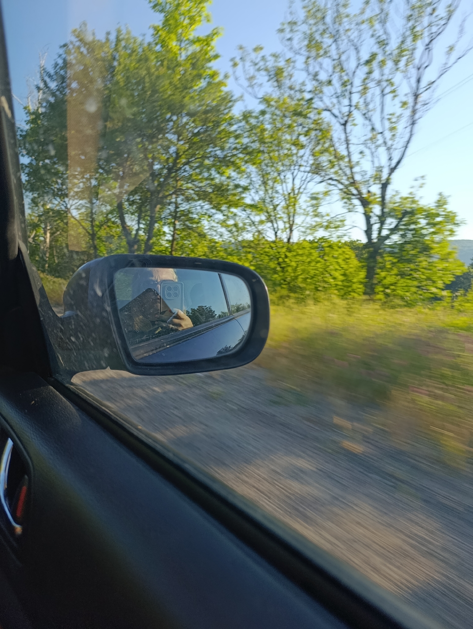 The image shows a reflection in a car's side mirror of a person holding a smartphone, taking a selfie while driving. The background reflects trees and a clear sky, suggesting an outdoor environment. The image also captures a portion of the car's interior, including the door panel and handle. The location is identified as Cherna skala, Bulgaria.