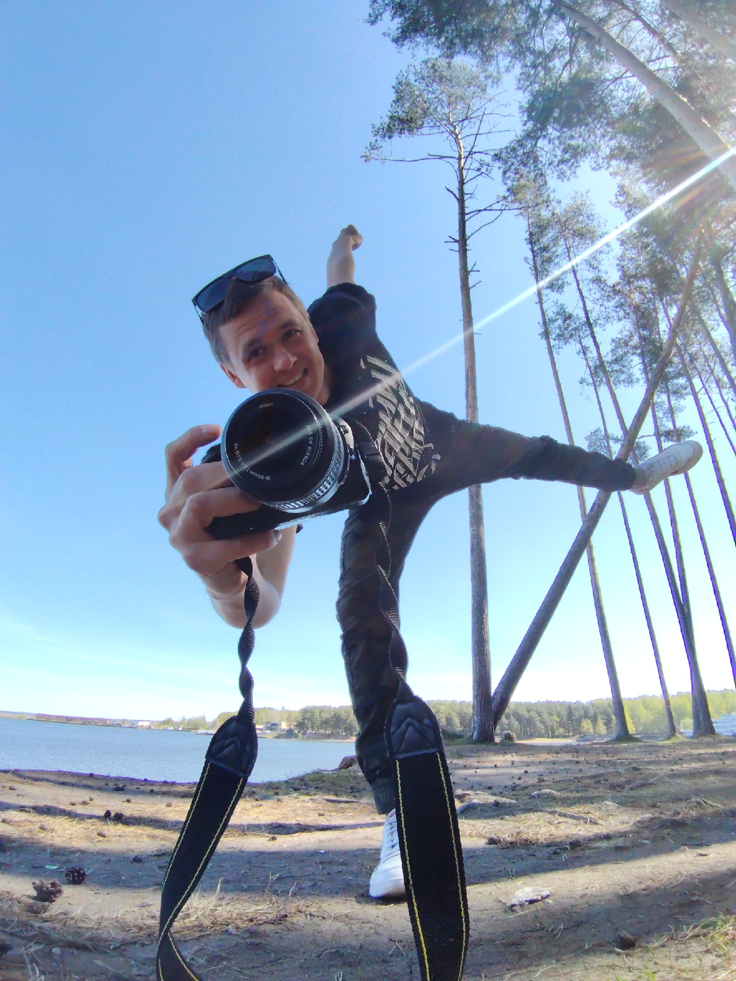 A man is posing in a forest clearing, holding a Nikon camera with a 18-55mm lens and sunglasses on his head. He has one leg extended out to the right, white shoes on his feet, and is standing on his left leg. He is wearing a black shirt and dark pants. The ground is dirt and pine needles. Tall pine trees surround the clearing, with a view of a lake and coastline in the distance under a blue sky. The location appears to be in the city of Chaikovsky, Russia.