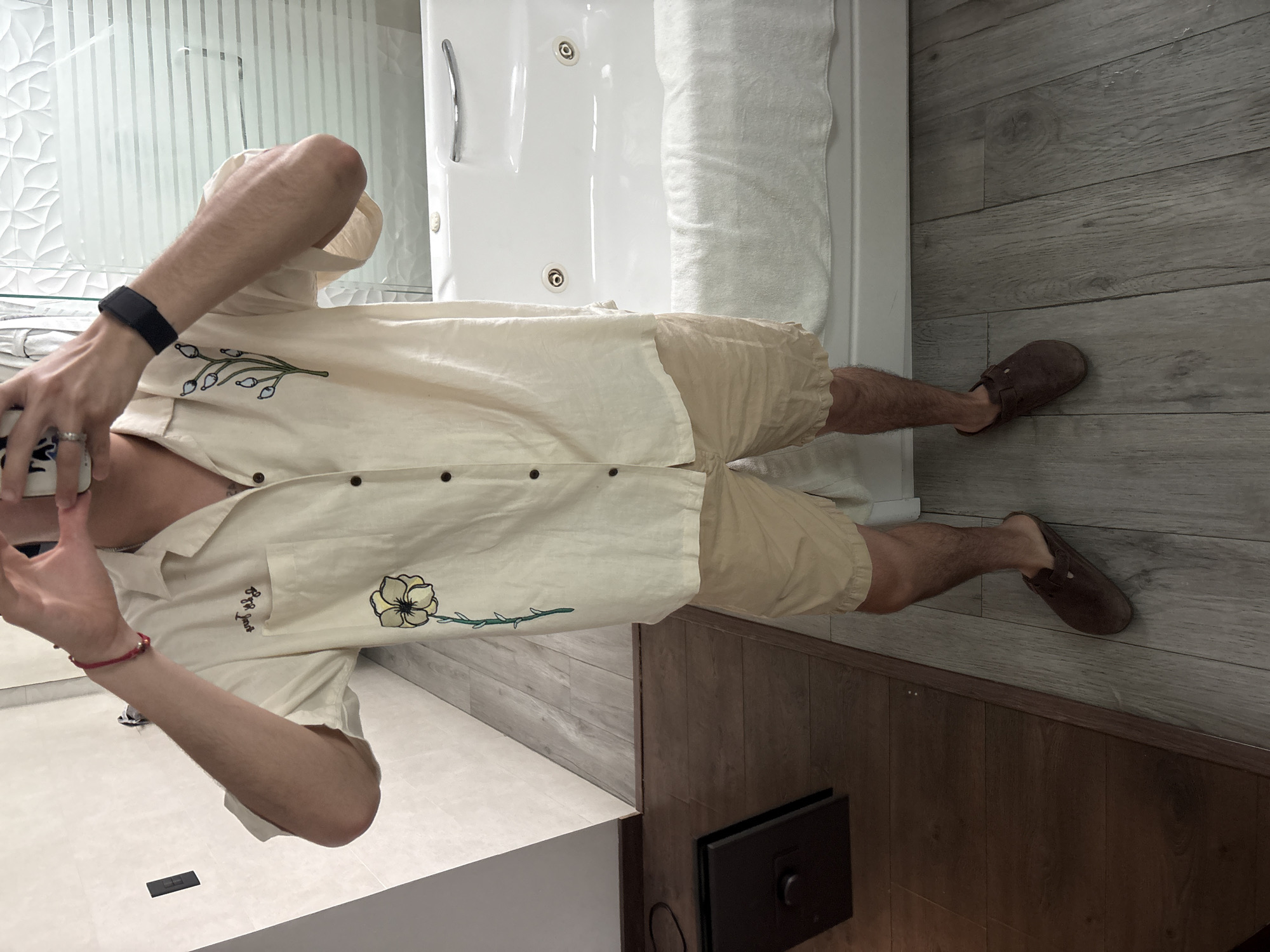 A person stands in a bathroom with one foot on gray wood-look tile and another on dark brown wood paneling. The person is wearing a tan shirt with floral embroidery, khaki shorts, and brown sandals. A white bathtub is partially visible. A black smart home device rests on the floor in the corner of the room. The location appears to be a private residence, as per the reference to San Bernardino Tlaxcalancingo, Mexico.