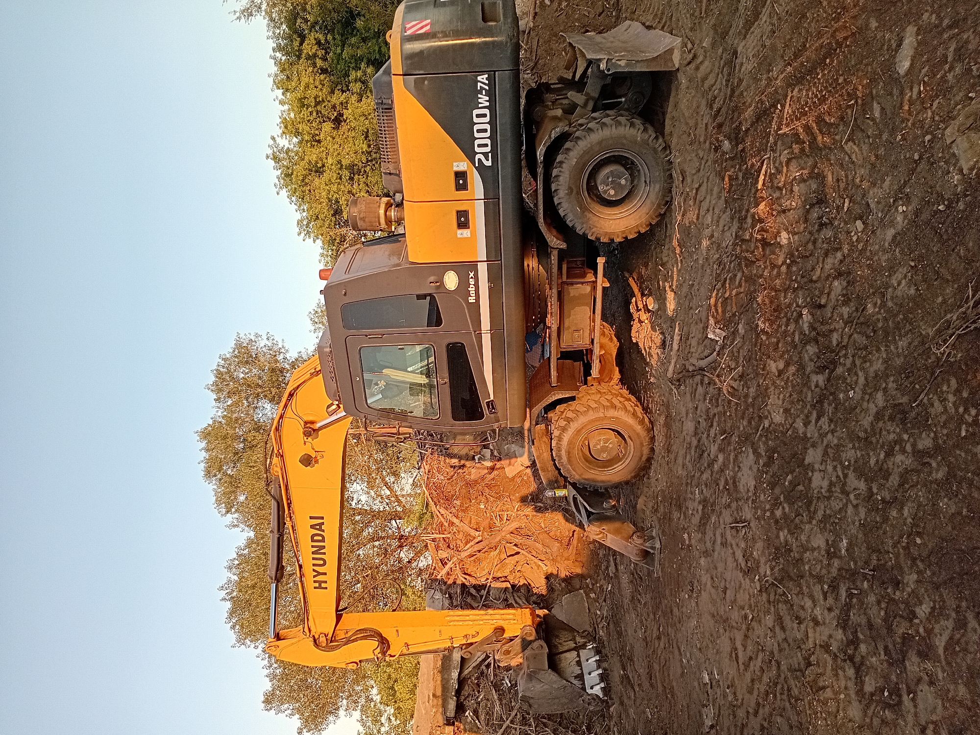 An excavator manufactured by Hyundai, model 2000W-7A, is parked on a dirt lot in Semey, Kazakhstan. The excavator is yellow and black and appears to be engaged in demolition or land clearing, as evidenced by a large pile of debris, including concrete and tree roots, near its bucket. The ground is muddy and uneven. Trees and foliage are visible in the background under a clear sky. The lighting suggests it is either early morning or late afternoon. No people are visible in the image.