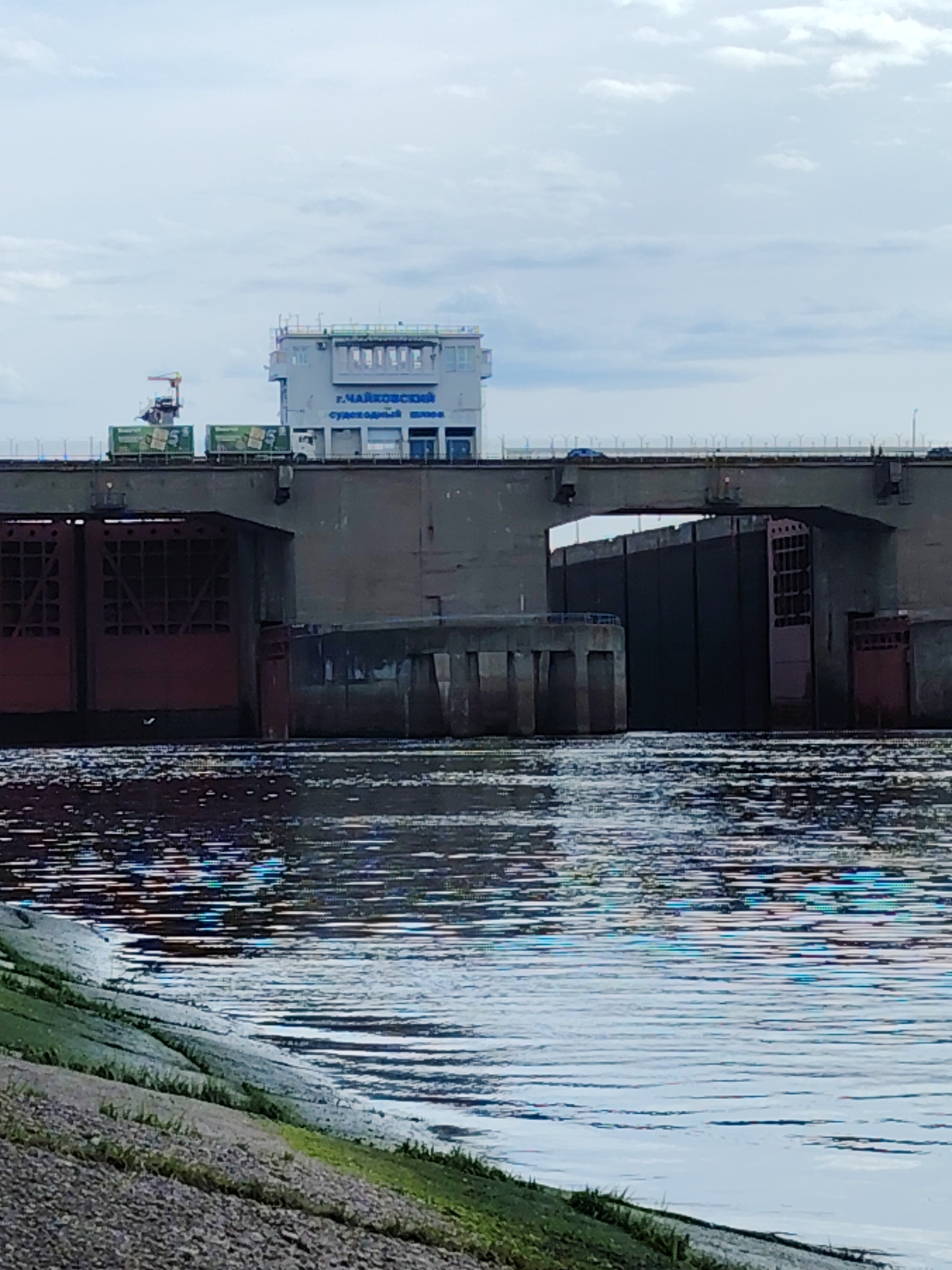 The image depicts the Chaikovsky Lock in the city of Chaikovsky, Russia. The lock structure consists of concrete and metal gates. The lock tower has signage in Russian. There's a bridge across the lock system, with vehicles visible on it. The water surface is rippled. The foreground shows a grassy riverbank. The sky is overcast.