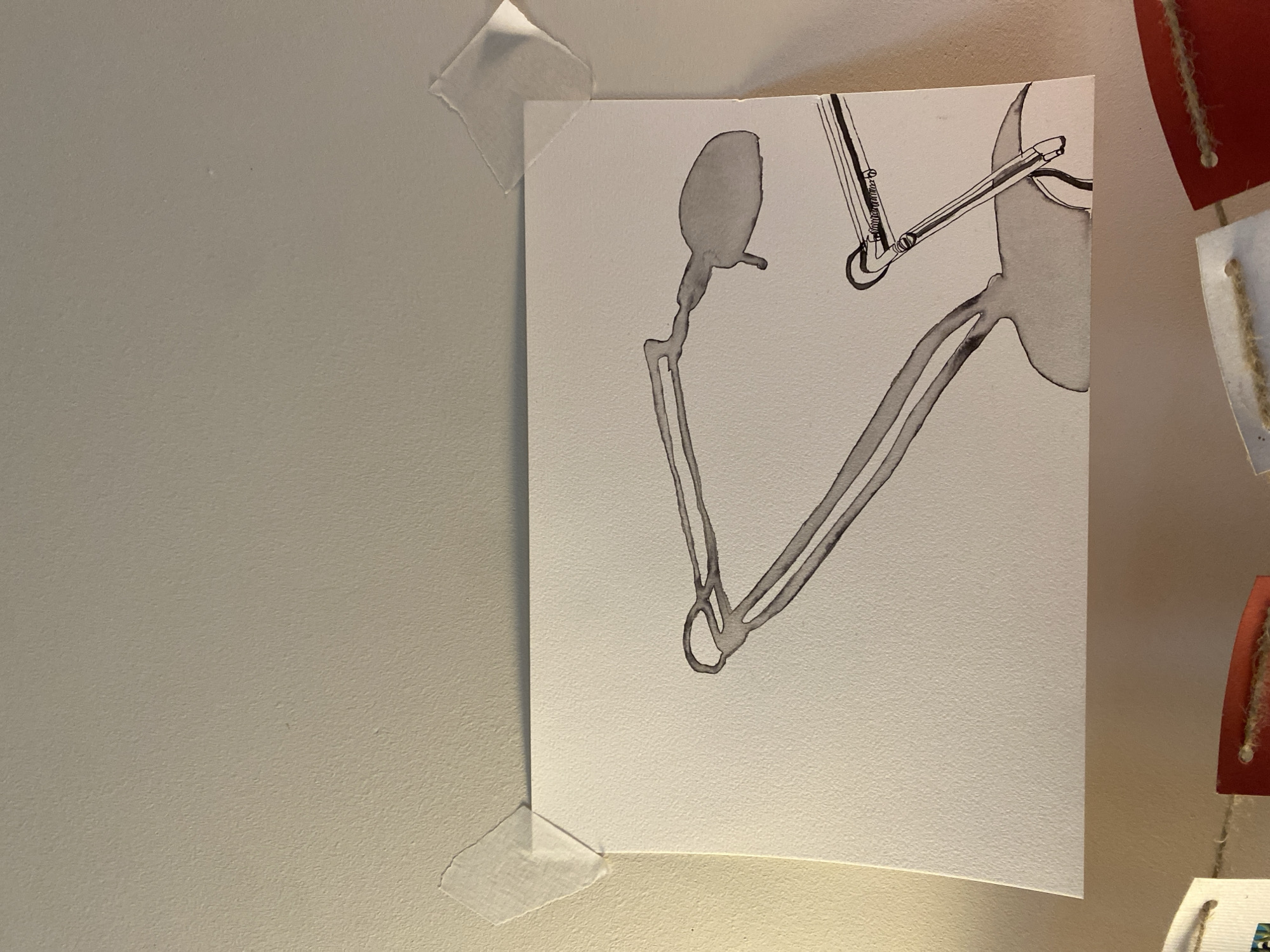 The image shows a drawing taped to a wall. The drawing is done in shades of gray and depicts abstract shapes and lines, possibly representing a figure or object. The wall is a light, neutral color. The drawing is held in place by two pieces of tape, one at the top left corner and another at the bottom left corner. To the right of the drawing, a decorative garland or hanging ornament with red and off-white elements can be partially seen. There is no information to indicate the city of Moscow, Russia.