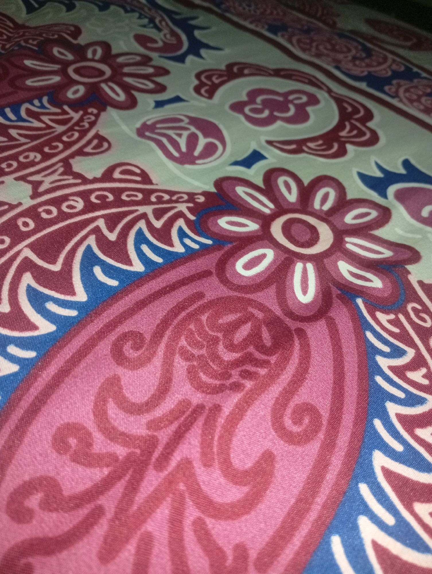 A close-up, high-angle shot captures a patterned textile, possibly a bedsheet or fabric, featuring a design with stylized floral and paisley motifs in shades of pink, maroon, and blue against a light-colored background. The fabric exhibits a slight sheen and visible texture. The image is devoid of any human presence, identifiable landmarks, or contextual environmental details that would indicate its specific location within Nyalindung, Indonesia. The scene is purely focused on the material's design and texture.