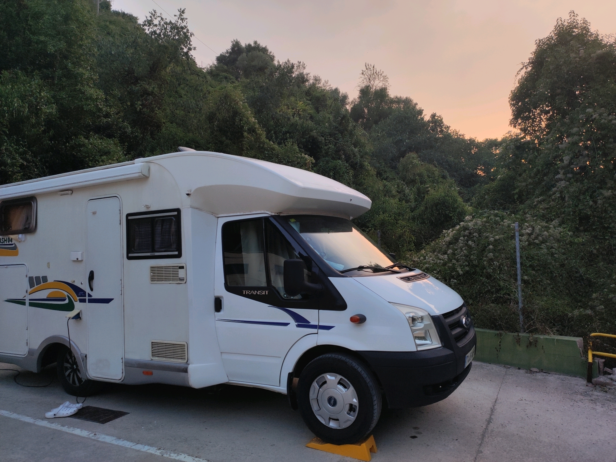 In Suera, Spain, a white motorhome is parked on a concrete surface next to a dense green hillside under a pale orange sky at dusk. The motorhome, a Ford Transit, has a white camper shell with blue and green decals on its side. A white cable is plugged into the side of the motorhome. A pair of white clogs rests on a doormat in front of the rear wheel. The front wheel of the motorhome is elevated by a yellow chocking block. The environment is natural, with lush vegetation covering the hillside and some faint power lines visible against the sky. The scene is still and quiet, suggesting a stop for the night or a brief rest.