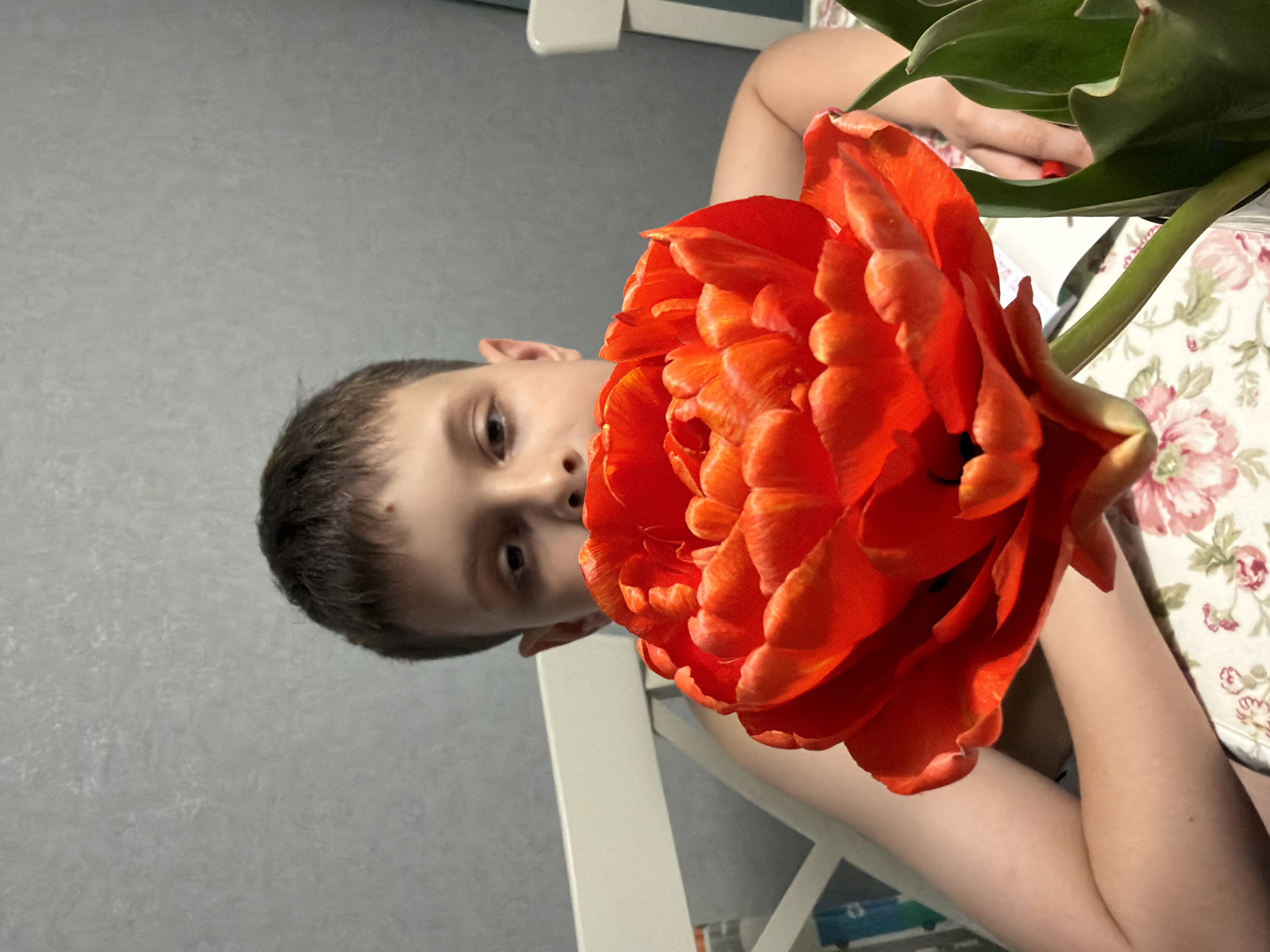 A boy with short brown hair is partially visible behind a large orange tulip flower. The boy's face is turned towards the viewer, with his eyes visible. The flower is in focus, its petals filling much of the frame. The background consists of a gray wall and a white chair. A floral-patterned surface, likely a tablecloth, is seen to the right. The photo appears to be taken indoors. No specific information about Votkinsk, Russia, is discernible from the image.