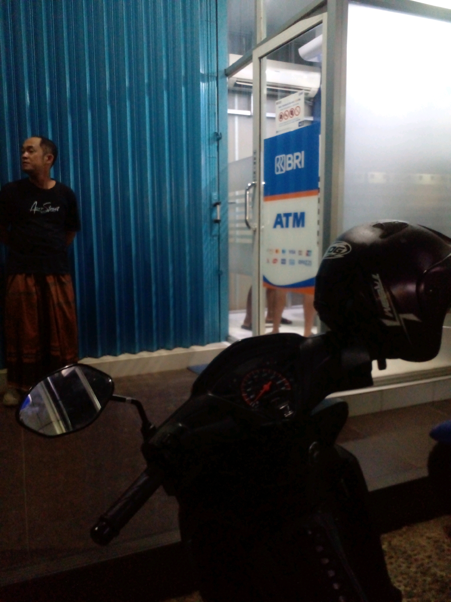 The image depicts an exterior scene at dusk, possibly in the city of Banyuputih, Indonesia. In the foreground, the handlebars and dashboard of a dark-colored motorcycle are visible, along with its side mirror reflecting a blurred image. A dark-colored helmet rests on the motorcycle's handlebars. Behind the motorcycle, a man stands facing left, wearing a black t-shirt with a white logo and brown patterned sarong. He is positioned in front of a bright blue corrugated metal roll-up door. To the right of the man and the blue door, an entrance to a building is visible. This entrance features a glass door with a silver frame and a sign indicating "BRI ATM". The ATM sign has a blue background with white text and a smaller orange stripe above it. The interior of the ATM area is dimly lit, and a white air conditioning unit is mounted on the wall. The ground appears to be paved with tiles. The overall lighting suggests it is either early morning or late evening.