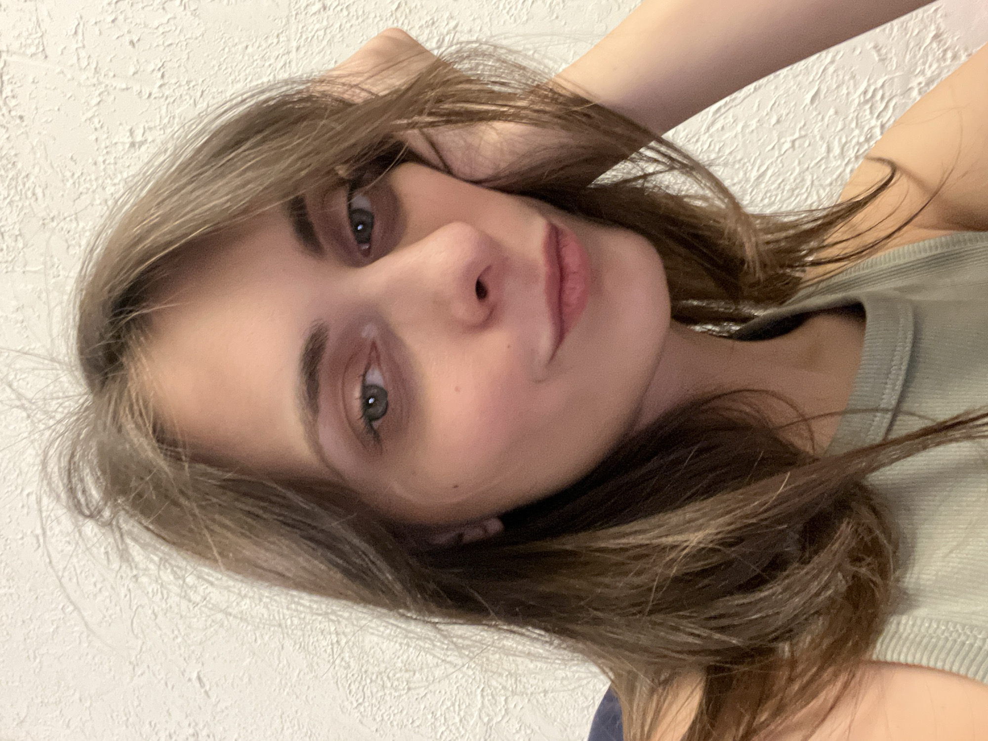 A close-up, rotated selfie of a fair-skinned woman with light brown hair. She is wearing a light green tank top. Her left hand is near her head, possibly touching her hair. The background is a textured white wall. There is no indication of Krasnoyarsk, Russia in the image.