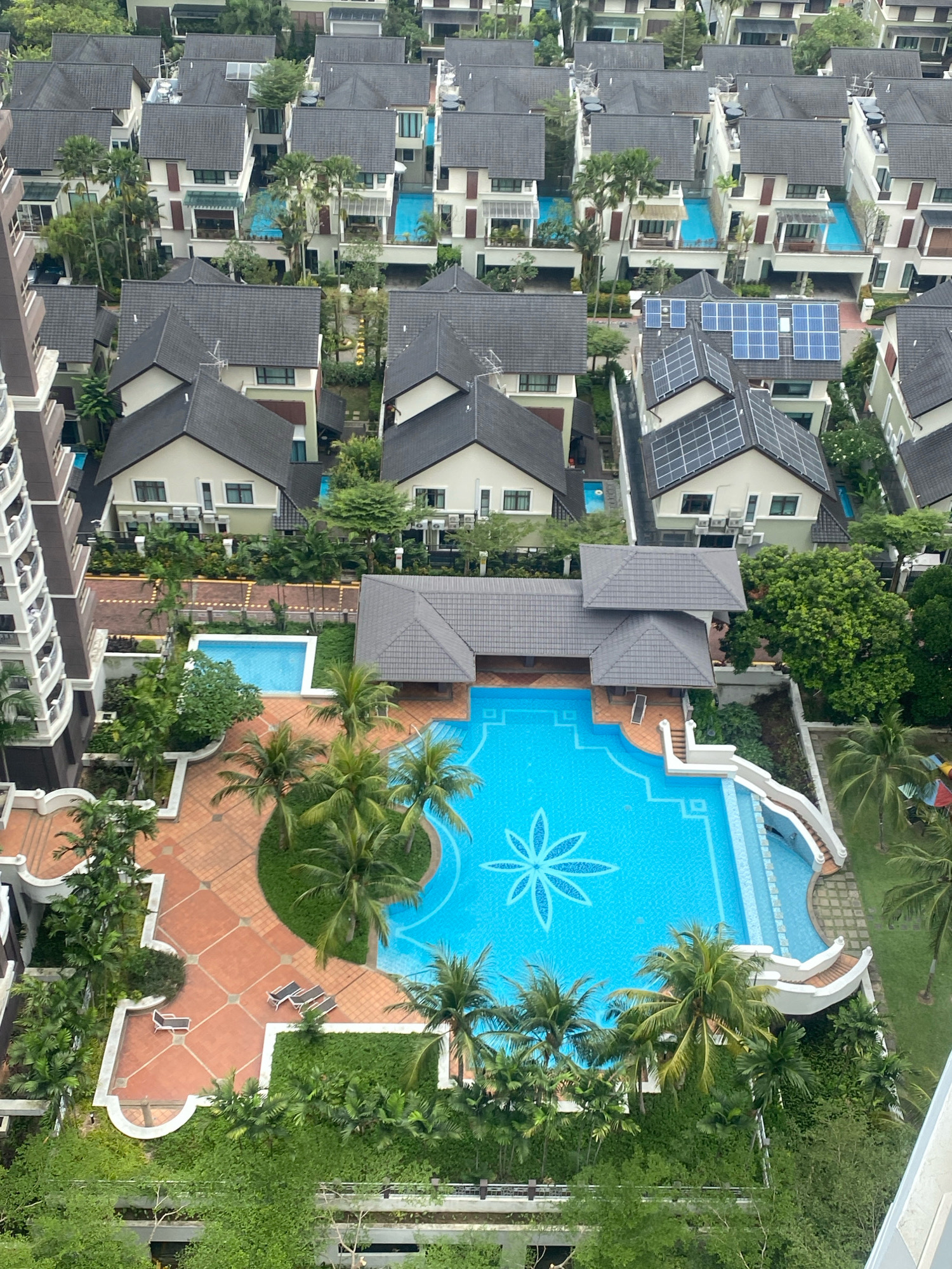 The aerial view shows a residential area in Kuala Lumpur, Malaysia, featuring a complex of modern housing with various amenities. In the foreground, a large, bright blue swimming pool with a decorative floral pattern in its center is prominently displayed. A white, winding slide enters the pool from the right, and a smaller rectangular pool is located to the left. The pool area is surrounded by red brick paving and lush green tropical landscaping, including numerous palm trees. Several lounge chairs are placed on the paving. Adjacent to the pool area are covered structures with grey tiled roofs, likely changing rooms or gazebos.
Beyond the pool area, a dense collection of multi-story houses with dark grey tiled roofs is visible. Many of these houses have private pools, indicated by patches of blue water within their courtyards. Some roofs are equipped with solar panels. The houses are modern in design, with white and cream-colored facades, large windows, and balconies. A tall, modern apartment building with a grey exterior is partially visible on the left edge of the frame, suggesting this residential complex is near or part of a larger urban development. The overall impression is one of a well-maintained, affluent residential neighborhood within a tropical urban environment. No people are visible in the scene.