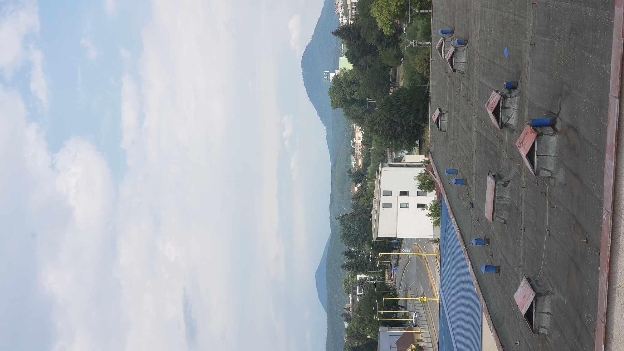 An elevated view of the city of Prešov, Slovakia, is depicted under a partly cloudy sky. In the foreground, a dark, weathered flat roof is visible, dotted with several ventilation pipes topped with blue caps, and various skylight structures with reddish-brown sloped coverings.
Beyond the roof, a modern white building with multiple windows stands out. To the left of this building, a road with yellow streetlights and traffic signs is present, with vehicles on it. Further into the background, lush green trees and smaller buildings form a dense urban landscape. In the distance, rolling hills covered in greenery, culminating in prominent mountain peaks, dominate the horizon. The overall scene suggests a typical urban environment on a summer day.