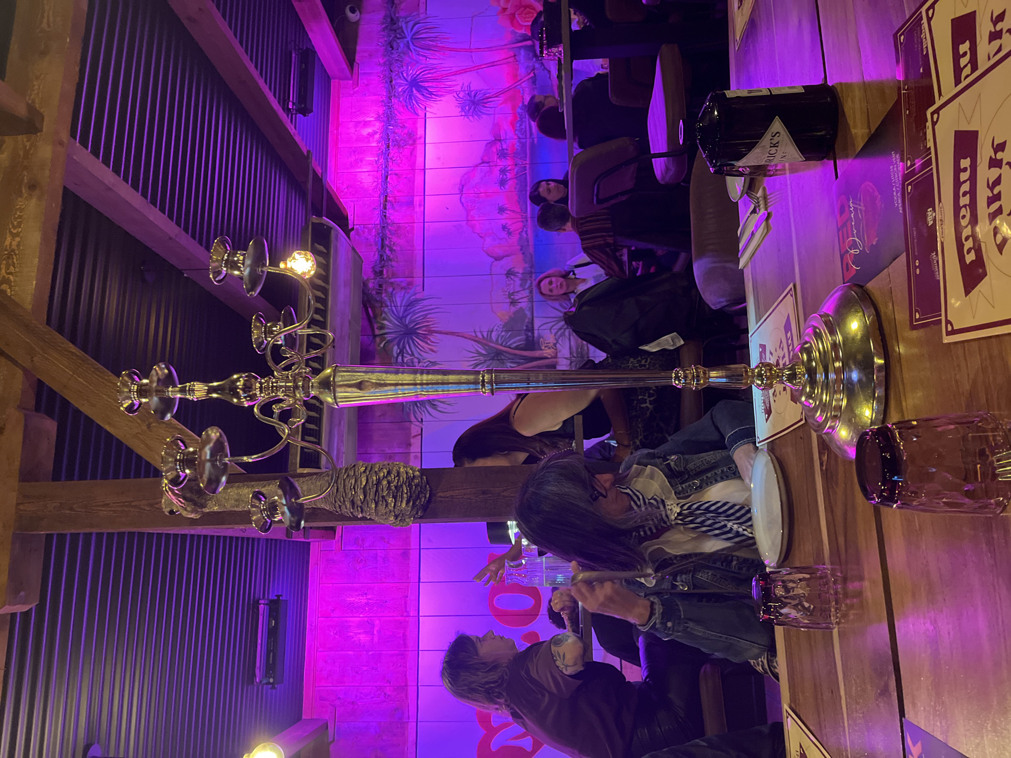 The image depicts the interior of the restaurant "ökkr" located in Saint-Jean-de-Védas, France. The scene shows a long wooden table with several people seated around it. On the table, there is a candelabra, plates, glasses, a bottle of Hendrick's gin, and menus. The wall behind the table is decorated with a mural featuring tropical plants and a pink and blue color scheme. The overall lighting is provided by pink and purple neon lights. The people appear to be dining and socializing.