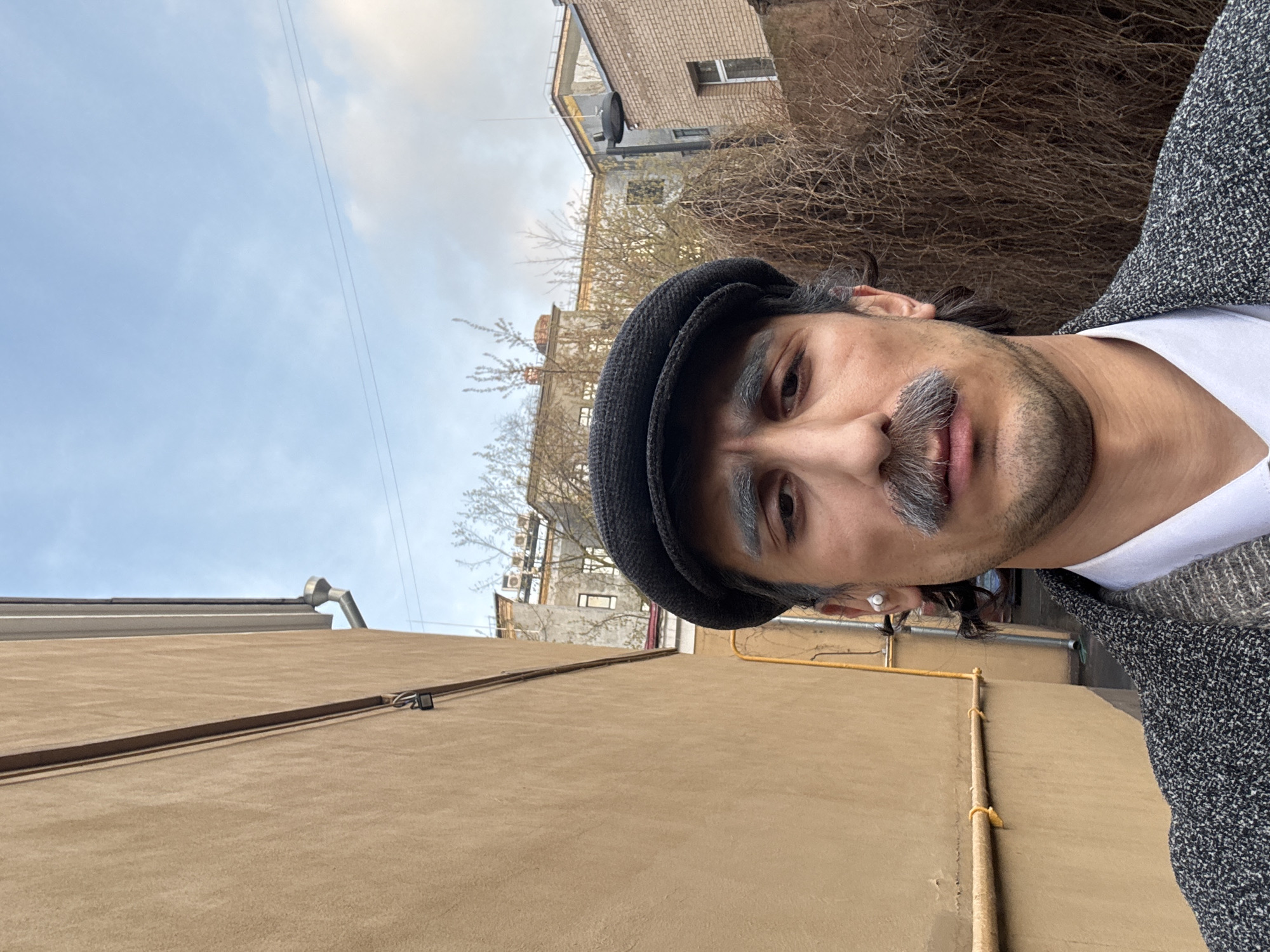 The image is a vertical close-up selfie of a man outdoors in an urban setting, likely Moscow, Russia. The man is positioned on the right side of the frame, looking directly at the camera with a neutral expression. He has dark hair, a distinct grey mustache, and a short, dark beard. He is wearing a dark, possibly tweed, flat cap, a white t-shirt, and a grey speckled cardigan or jacket. A small silver stud earring is visible in his left ear.
The background reveals various older buildings, some with exposed brick and others painted, under a light blue sky with some cloud cover. Bare tree branches or bushes are visible behind and to the left of the man, suggesting late autumn, winter, or early spring. A large, plain tan-colored wall occupies the lower-left portion of the frame, with some horizontal and vertical cables or pipes attached. Additional yellow pipes are also visible on walls in the mid-ground.
The time of day is daytime, with soft, diffused light, indicating either an overcast sky or an early morning/late afternoon hour without harsh shadows. The buildings and exposed utility lines contribute to an aged, urban aesthetic. There is no visible text in the image.