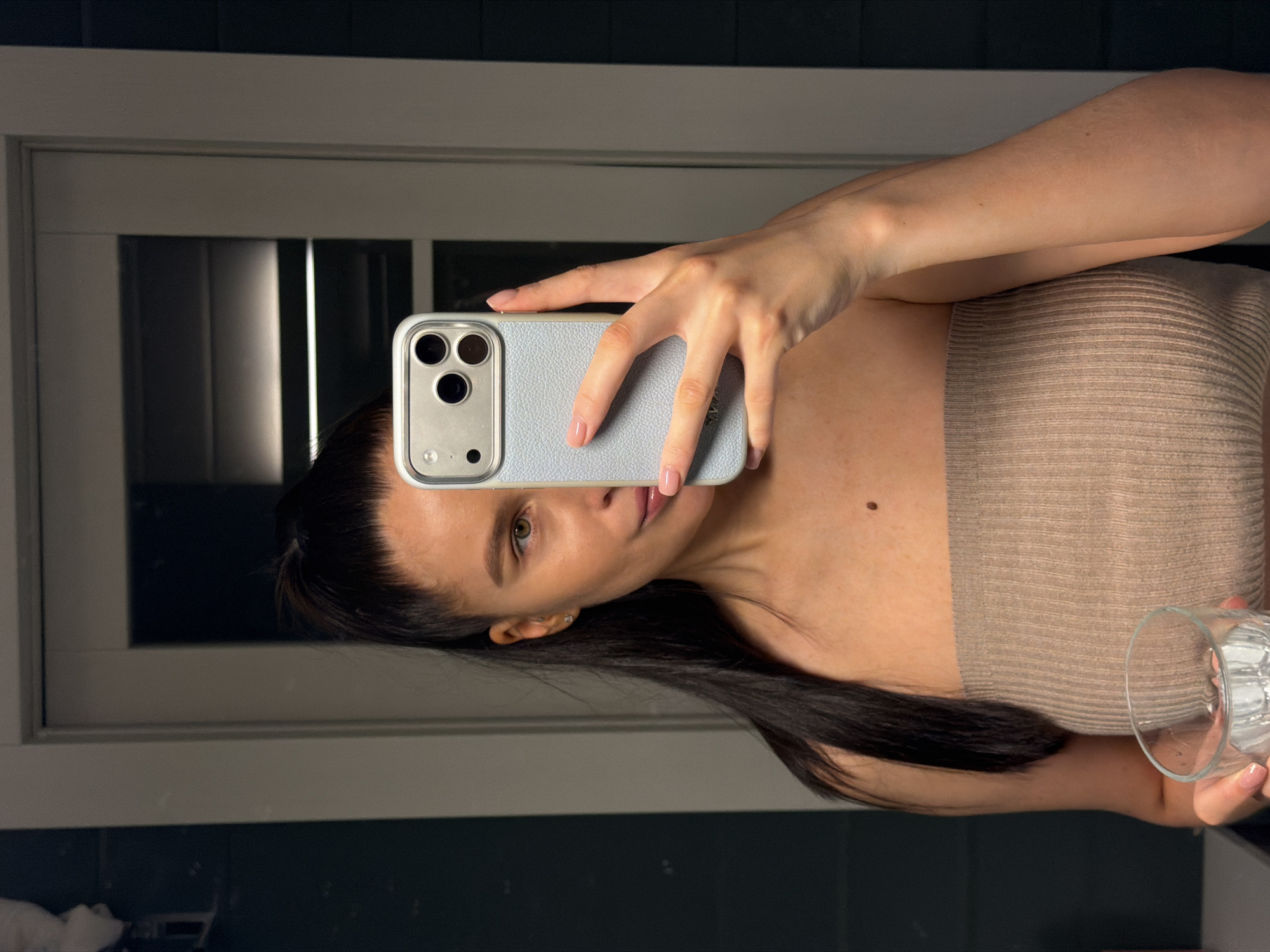 A woman is taking a selfie in a bathroom mirror. She is wearing a tan tube top. She is holding a glass of water in her right hand and a cell phone in her left hand. The bathroom has dark blue walls. The location is Unknown, Unknown.