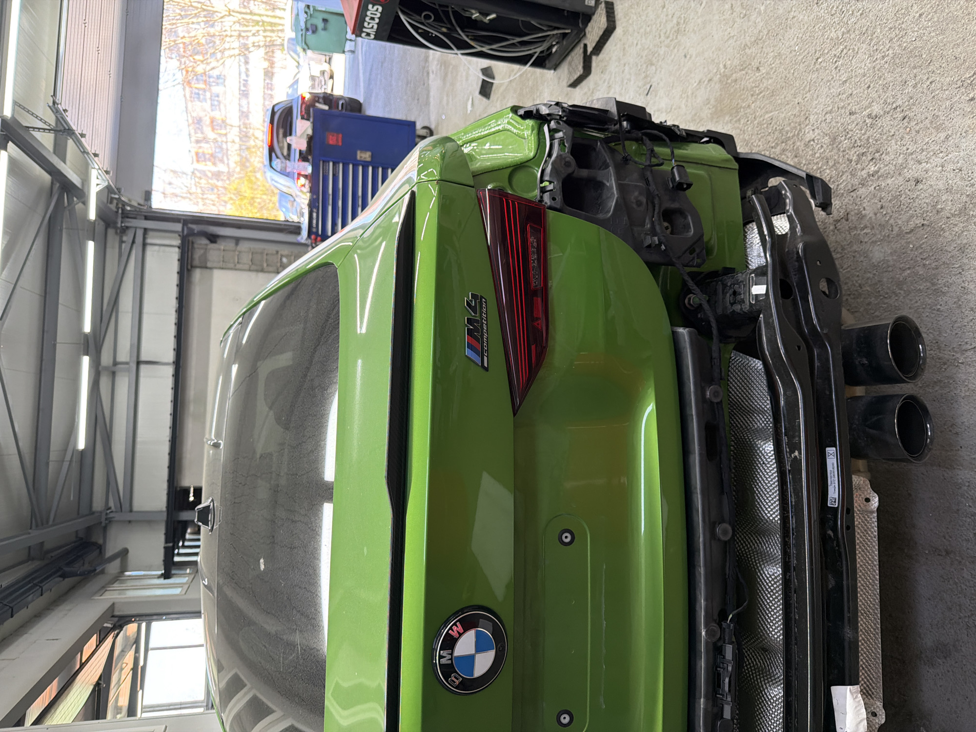 A bright green BMW M4 Competition, with its rear bumper removed, is the central subject in an automotive workshop. The car is oriented with its rear facing the viewer, slightly angled, revealing the internal structure of the bumper mounting points, a heat shield, and two black exhaust tips on the right side. The car's tailgate features a BMW emblem and an "M4 Competition" badge with the signature BMW M colors. The right rear taillight is illuminated.
The setting is a well-lit garage or body shop during the daytime, evidenced by natural light filtering through large windows or a roll-up door in the background, complemented by overhead fluorescent lighting. Various workshop equipment is visible, including a blue toolbox (partially showing a "Snap-on" logo) and a green piece of equipment labeled "CASCOS" in the background. The workshop walls appear to be made of corrugated metal, and a concrete floor is visible. The overall scene suggests the car is undergoing repair, modification, or maintenance. A white sticker with a barcode and text including "7 0 2 0", "2023 03 09", and "CE" is visible near the exhaust system.