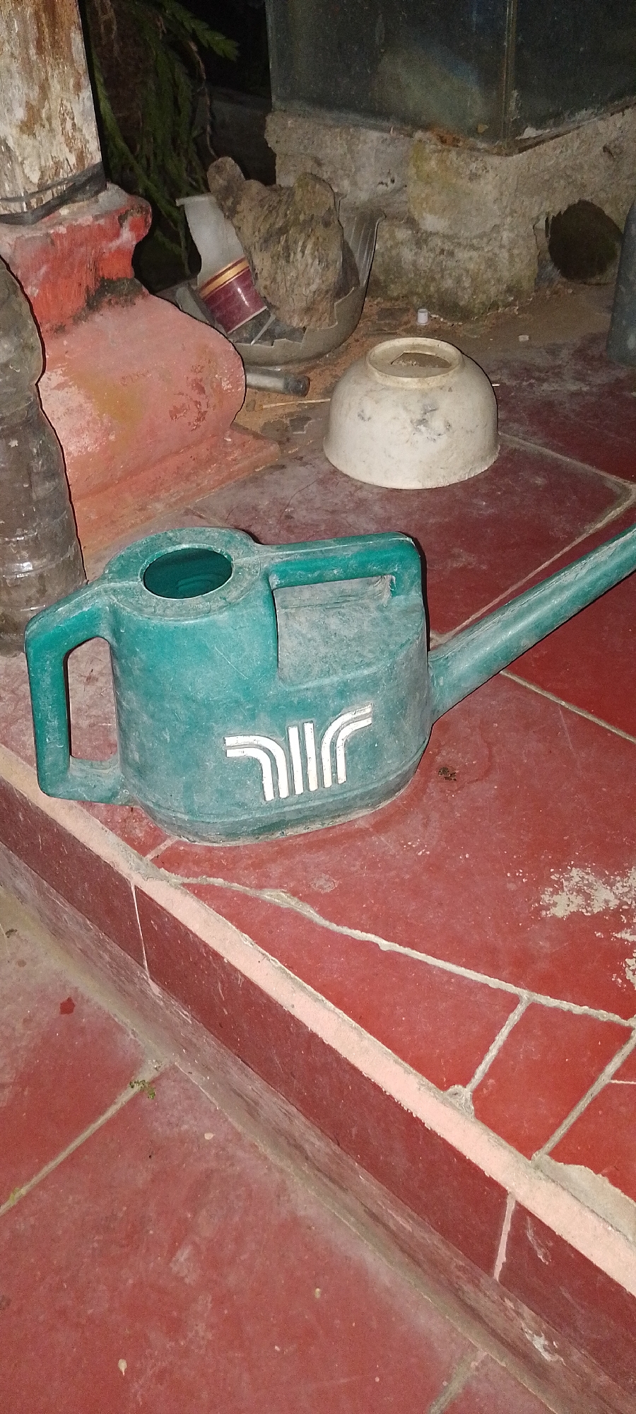 In Pekutatan, Indonesia, a teal watering can sits on a tiled surface. The watering can has a handle on top and a spout extending to the right. A white logo with vertical lines is visible on the side of the watering can. The tiled surface is red and appears to be outdoors, with some dirt and debris present. In the background, there are various objects, including a broken bowl, a bottle, and a portion of what appears to be a wall or structure. The lighting suggests it is nighttime or an indoor dimly lit area.