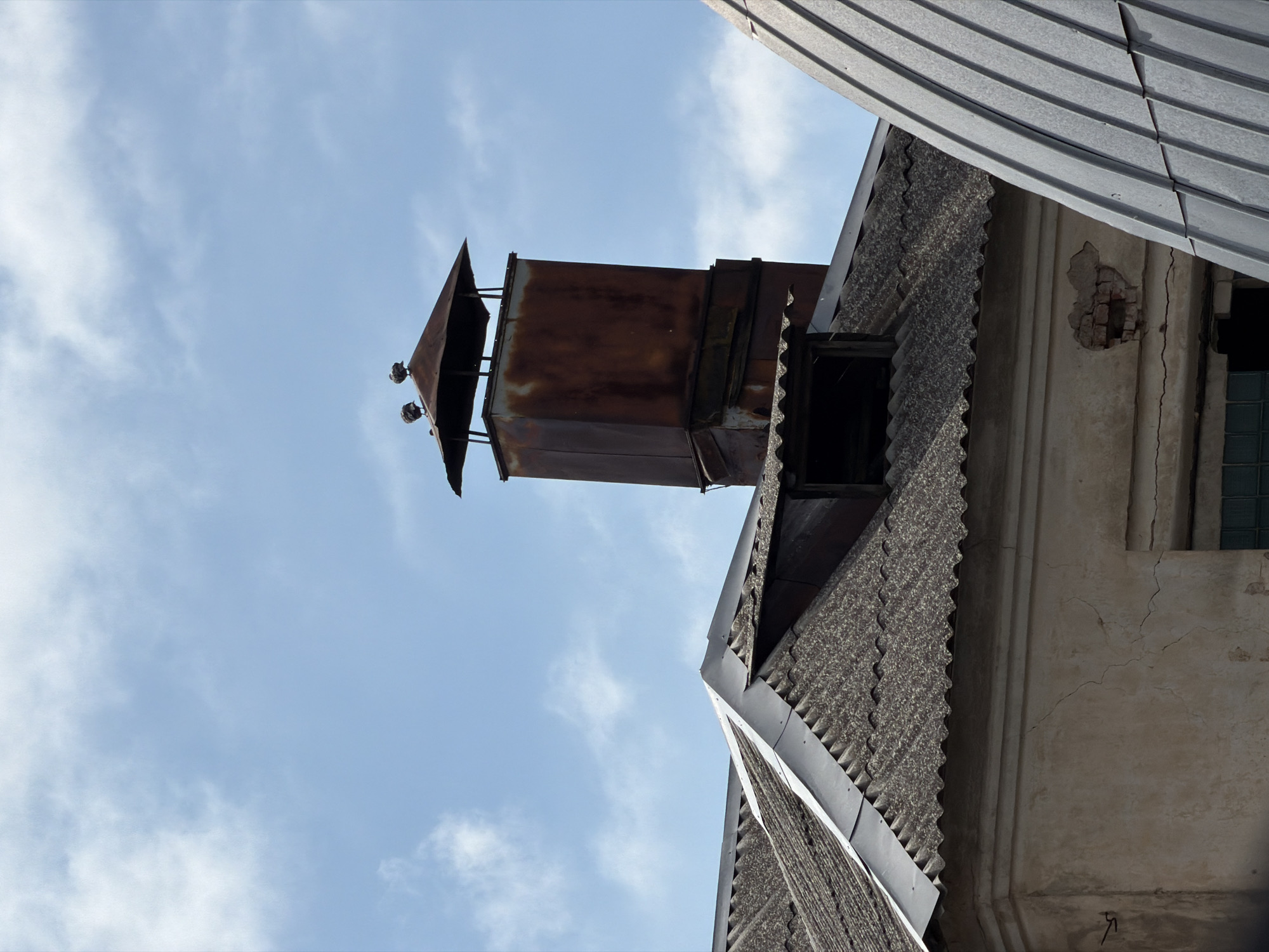 The image shows a close-up view of a rusted metal chimney with a square cap on a sloped roof, against a blue sky with scattered white clouds. Two birds are perched on top of the chimney cap. The roof is made of corrugated material, likely asbestos-cement shingles, and appears to be weathered. A portion of a pale-colored building with visible cracks in its stucco exterior is visible below the roofline. To the right of the chimney, a section of a building with a ribbed metal roof is also visible. The scene suggests an outdoor setting, likely in an urban or semi-urban area, possibly in Бягомльскі сельскі Савет, Belarus, given the provided context. There are no people visible in the image.