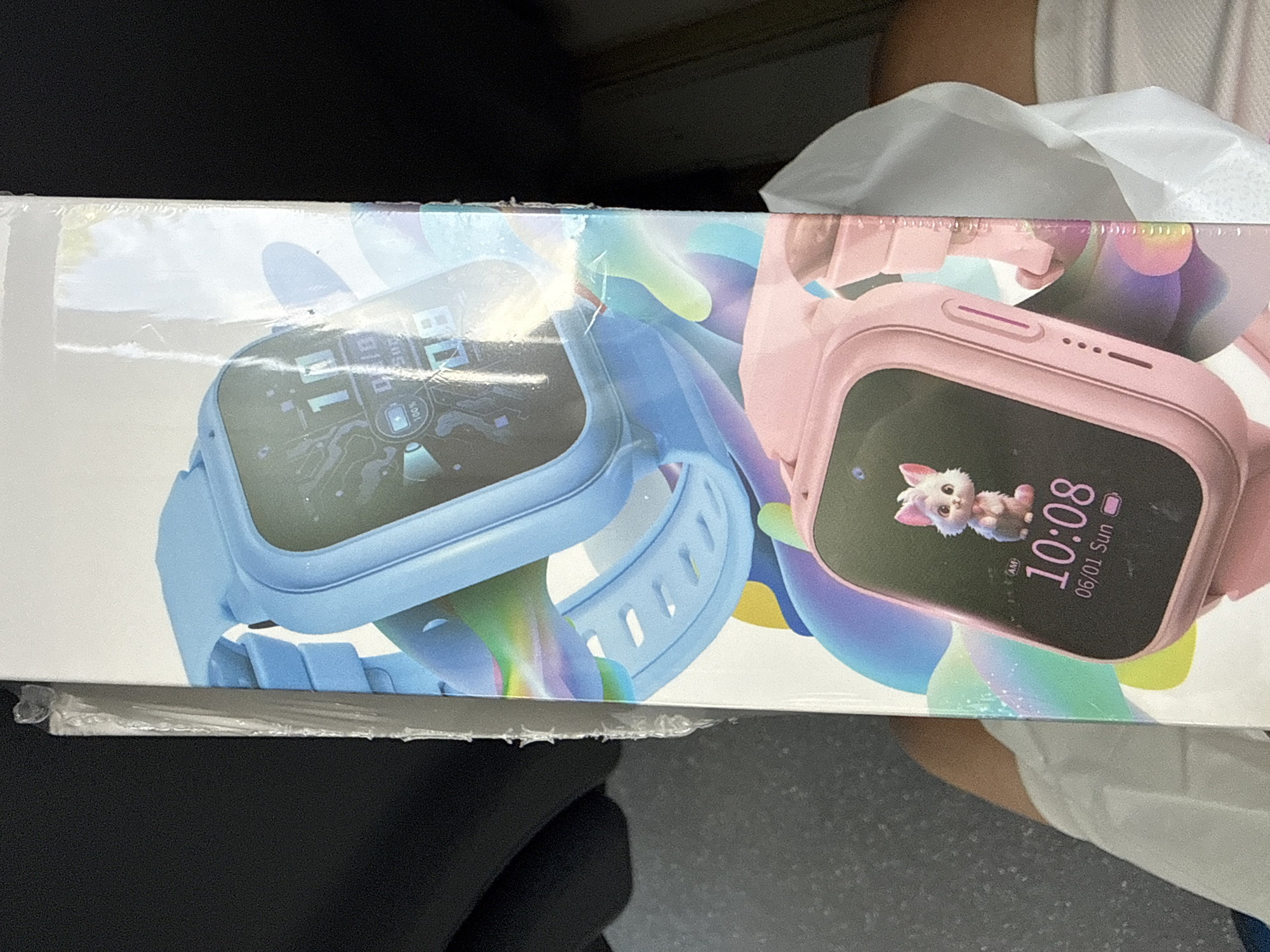 The image shows a close-up, eye-level view of a product box for children's smartwatches. The box is predominantly white with colorful graphics featuring two smartwatches, one blue and one pink. The blue smartwatch is shown at the top, displaying a digital interface with the time and what appears to be battery and signal indicators. The pink smartwatch is positioned below, also showing a digital display with the time "10:08", the date "06/01 Sun", and a cute cartoon cat icon. The box has a clear plastic shrink wrap covering it, which is slightly torn in places along the edge, particularly on the right side. The background is dark and out of focus, suggesting the photograph was taken indoors, potentially in a vehicle, given the slight curvature and lighting in the top left corner.