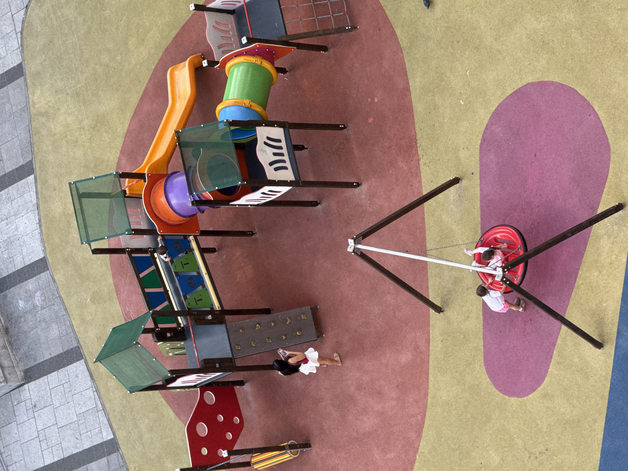 A high-angle shot depicts a colorful playground in an urban area, possibly Unknown, Unknown. The playground features multiple play structures, including a slide, climbing apparatus, and a spinning apparatus with two children inside. A woman wearing a white skirt lies near the play structures. The ground is covered in various colors of soft, rubber-like material. The playground is adjacent to a paved area.