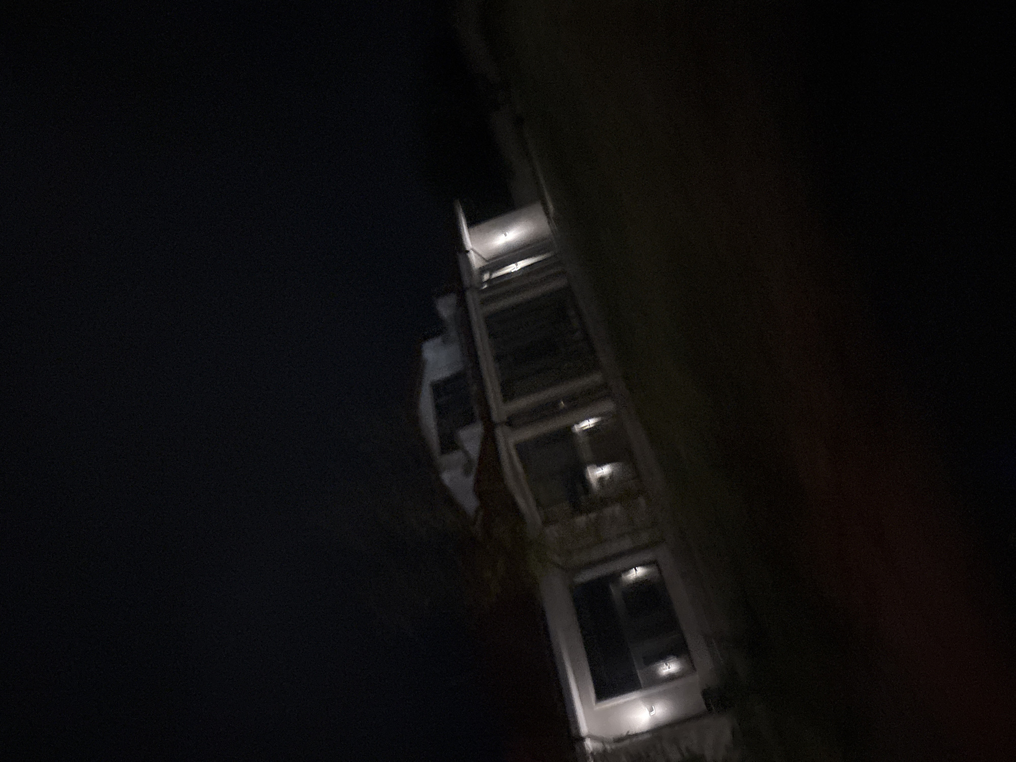 This media file captures a multi-story building at night, partially illuminated against a dark background. The scene is dominated by the upper and middle sections of the building, which features several windows, balconies, or terraces, and distinct architectural elements. Multiple light fixtures are visible, casting bright, localized illumination on the building's light-colored facade, likely concrete or stucco.
The image is taken from an angled perspective, possibly looking upwards or sideways at the structure, creating a dynamic and somewhat abstract composition. The surrounding area is largely obscured by darkness, with only faint outlines of what might be adjacent structures or foliage visible on the right edge. No people are visible, nor is there any discernible activity taking place. The time of day is clearly night, as indicated by the artificial lighting against the deep black sky. There are no specific weather cues, and no visible text can be identified. The location context suggests this building is in Plovdiv, Bulgaria, and its modern design is typical of contemporary urban architecture.